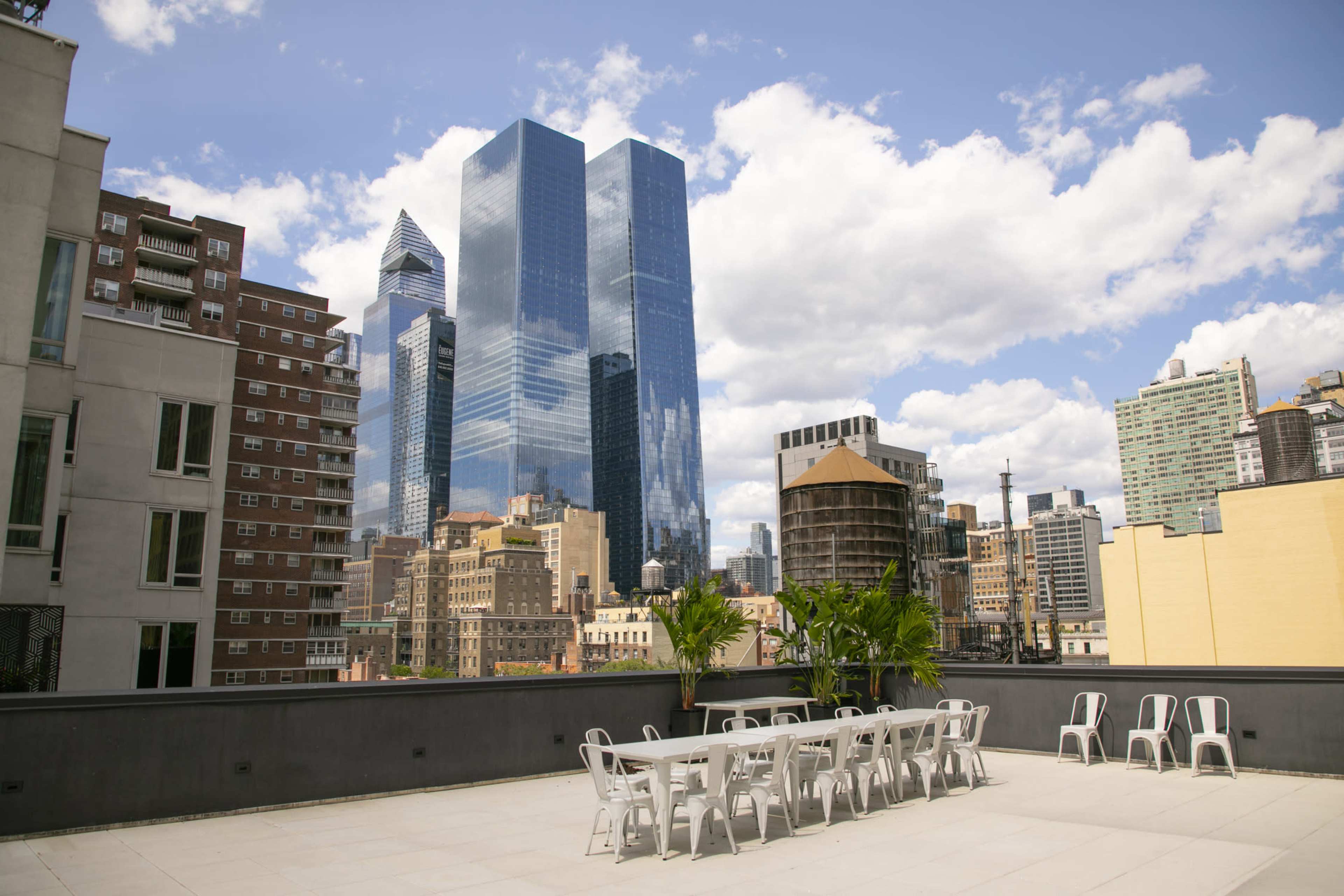 A rooftop terrace with a long table and chairs overlooks a skyline of modern skyscrapers under a partly cloudy sky.