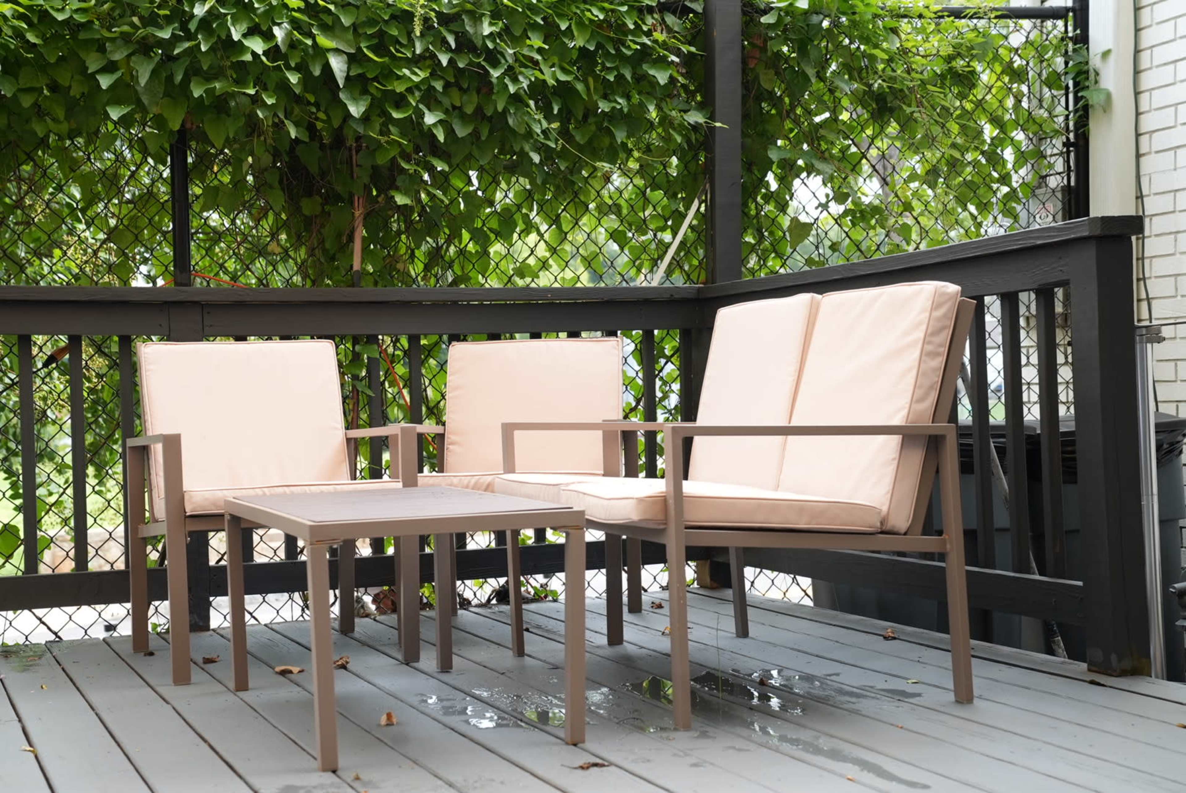 A set of light-colored outdoor furniture with two chairs and a small table is arranged on a wooden deck surrounded by lush greenery.