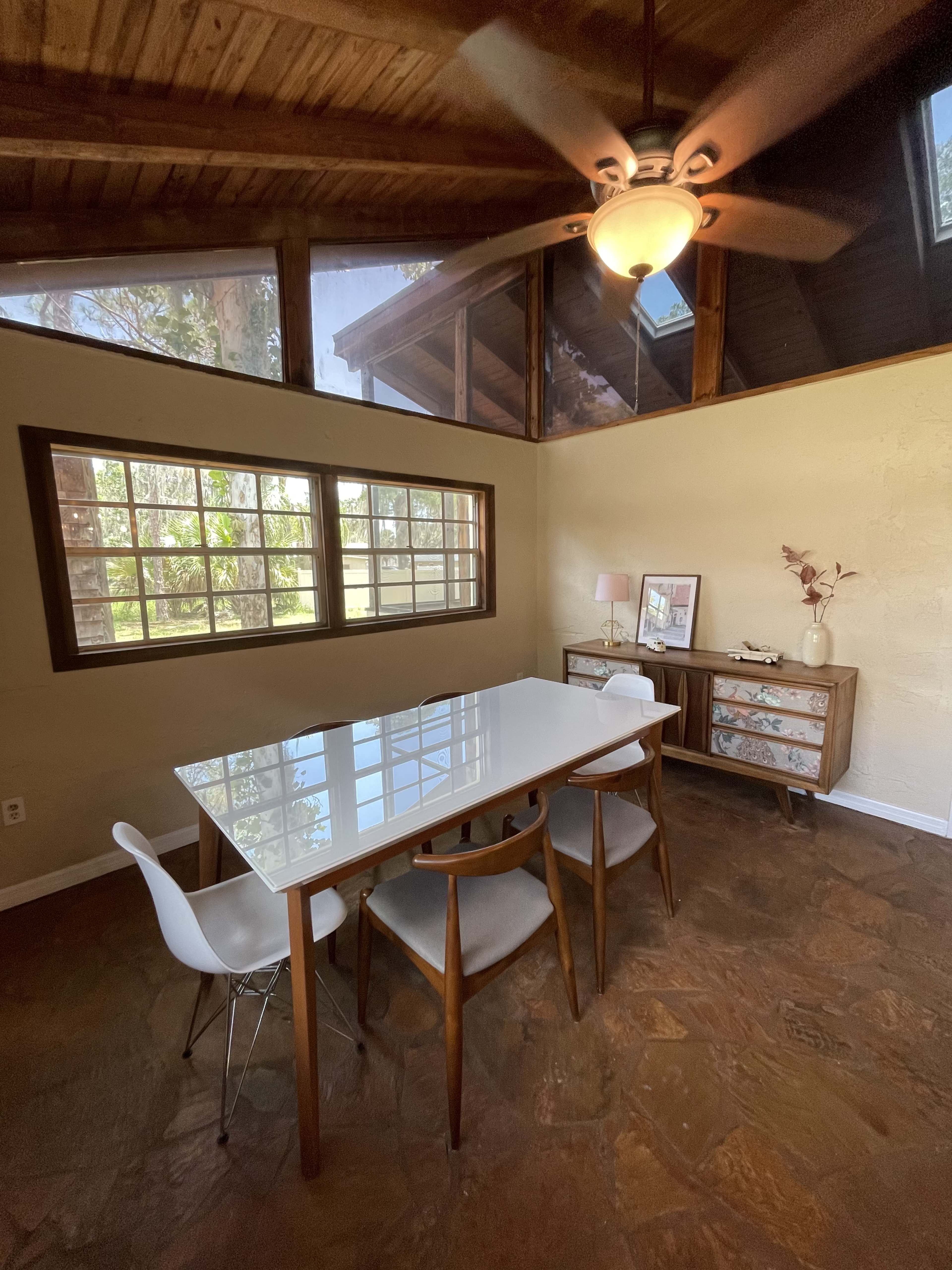 A modern dining area features a white table surrounded by wooden chairs, with a sideboard and large windows that provide natural light.