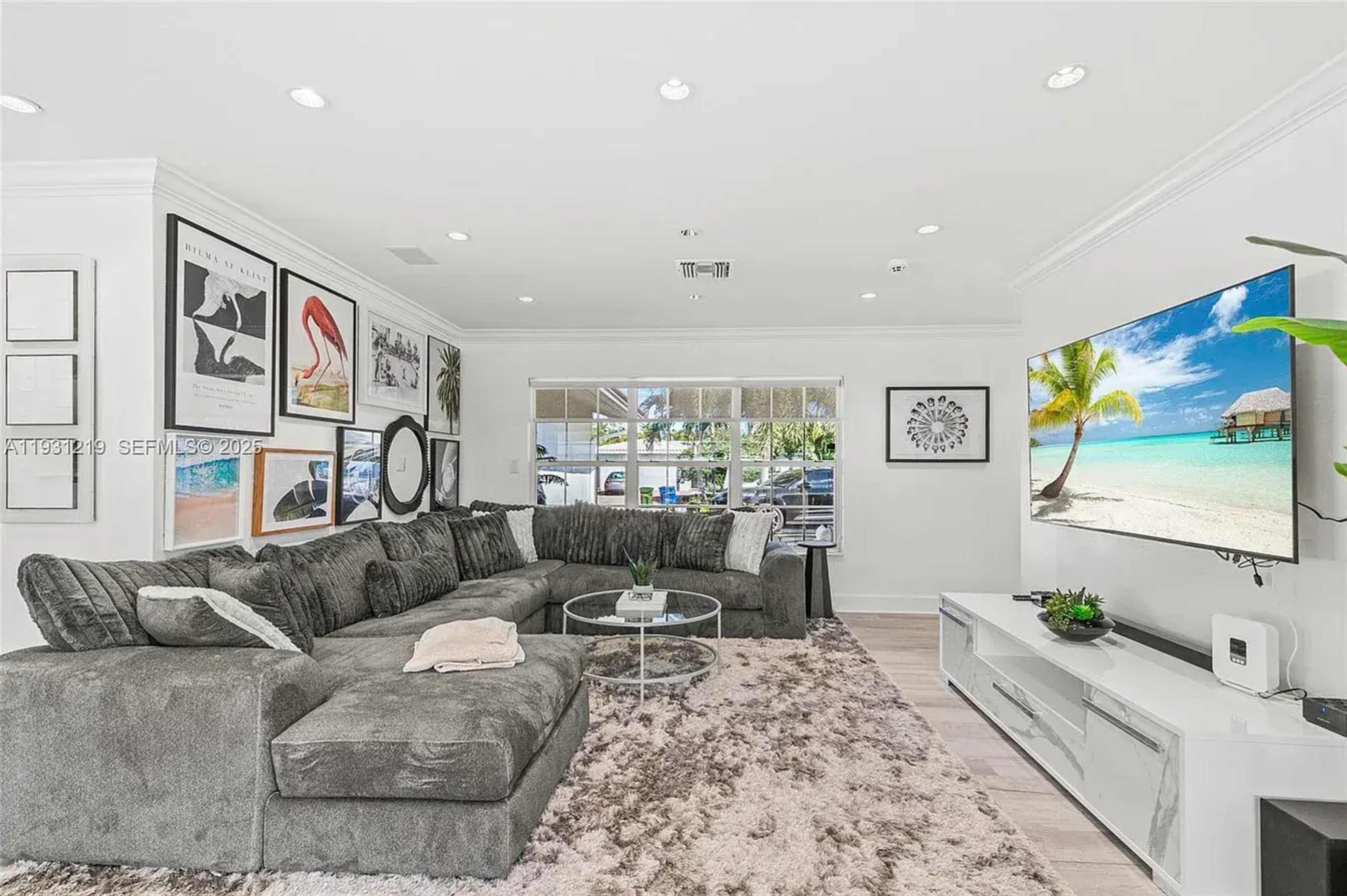 The image shows a modern living room with a large sectional sofa, a round glass coffee table, and a large flat-screen TV mounted on the wall.