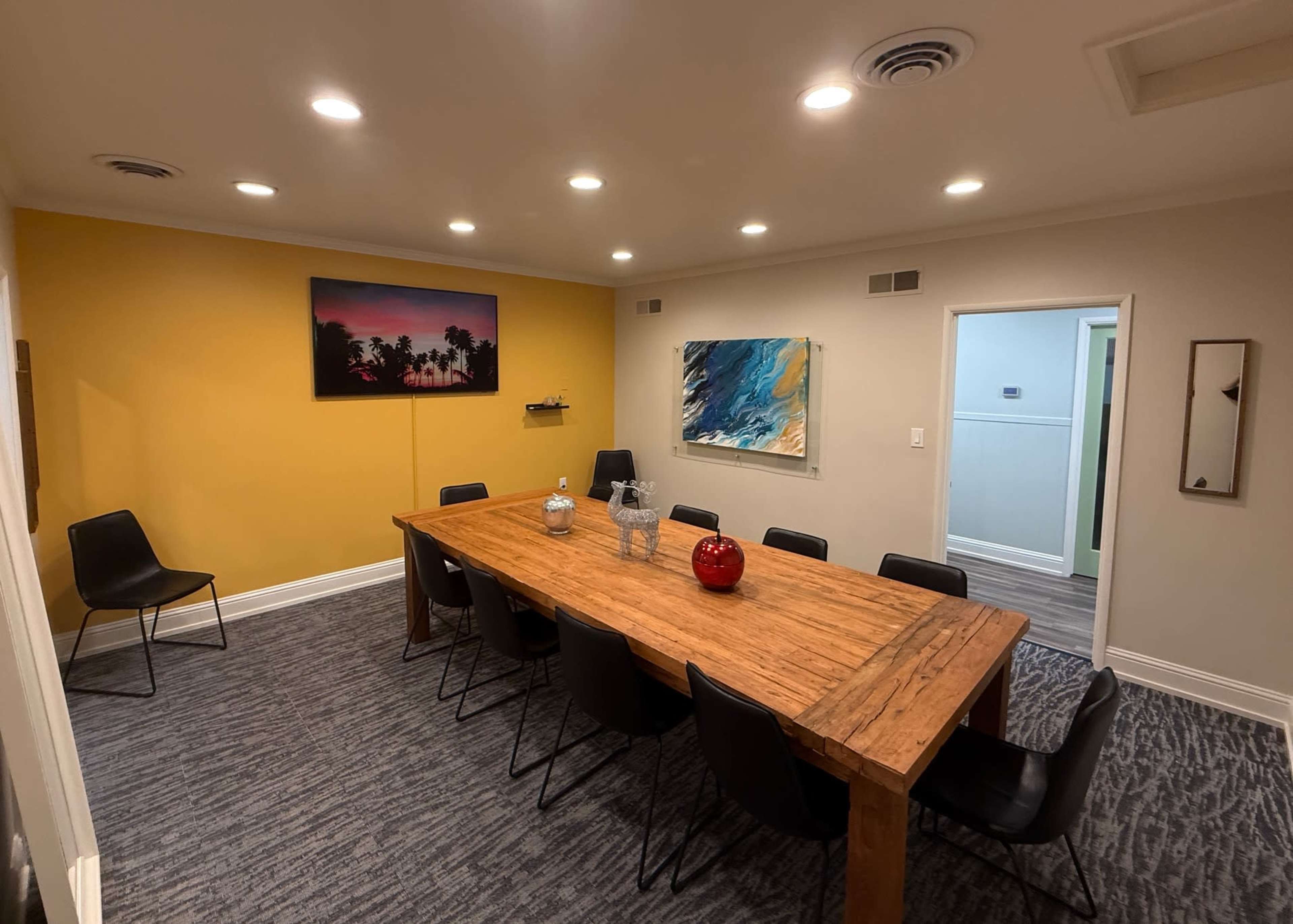 A long wooden table is surrounded by black chairs in a well-lit room featuring colorful paintings on the walls.