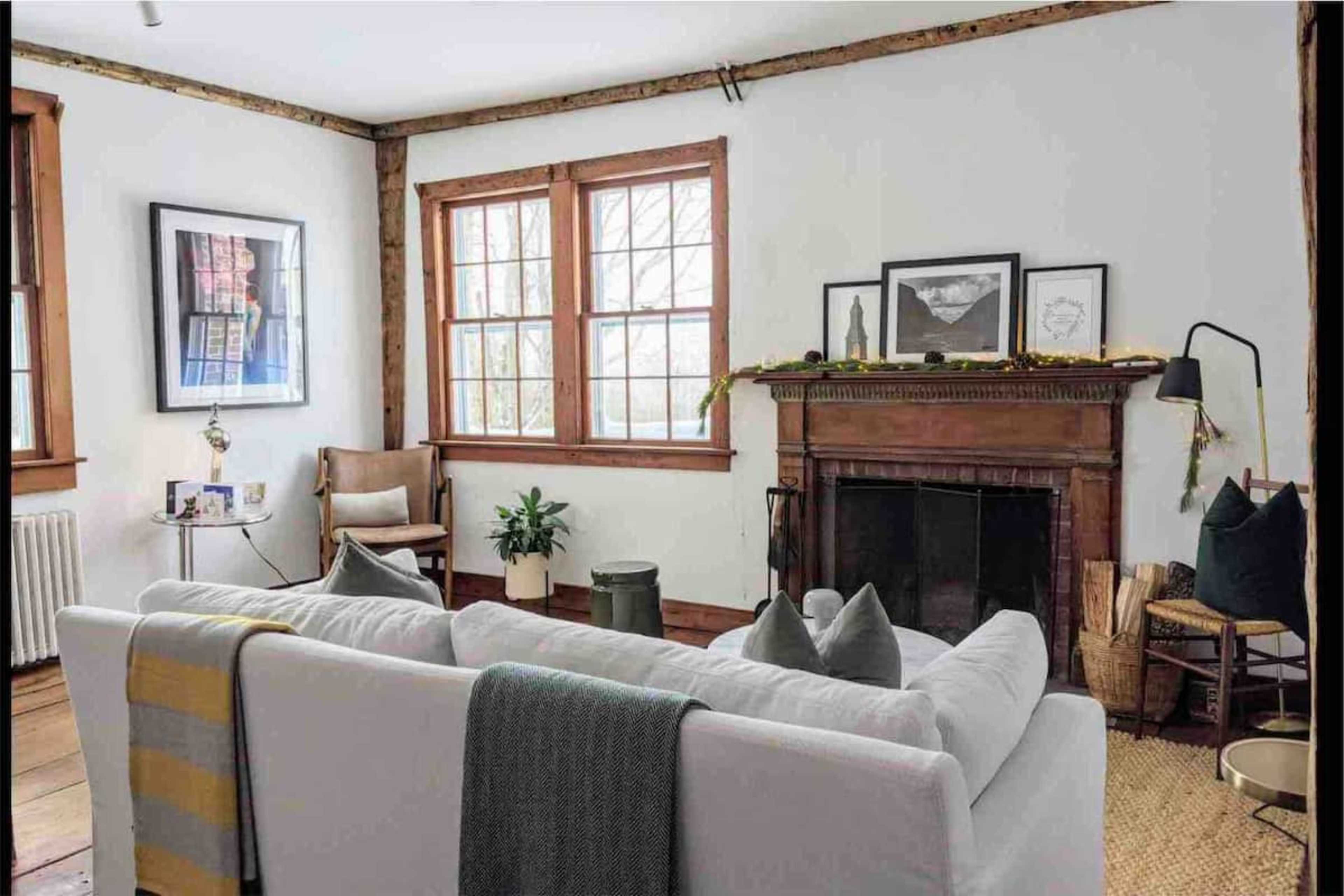 The image shows a cozy living room featuring a white sofa with a gray and yellow throw blanket, a wooden fireplace with decorative items, and large windows that allow natural light to enter the space.