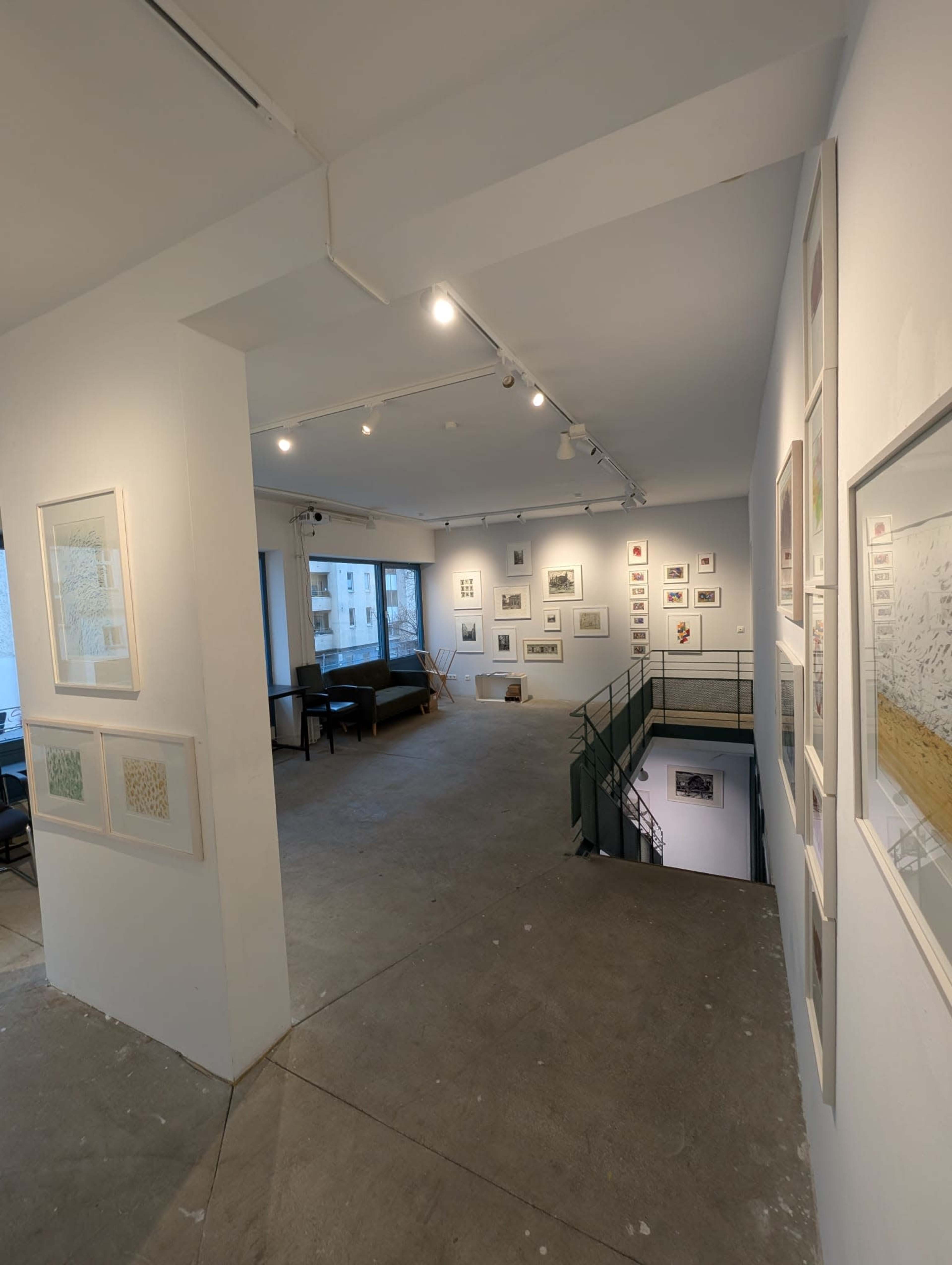 The image shows the interior of an art gallery featuring framed artworks displayed on white walls and a seating area in the background.