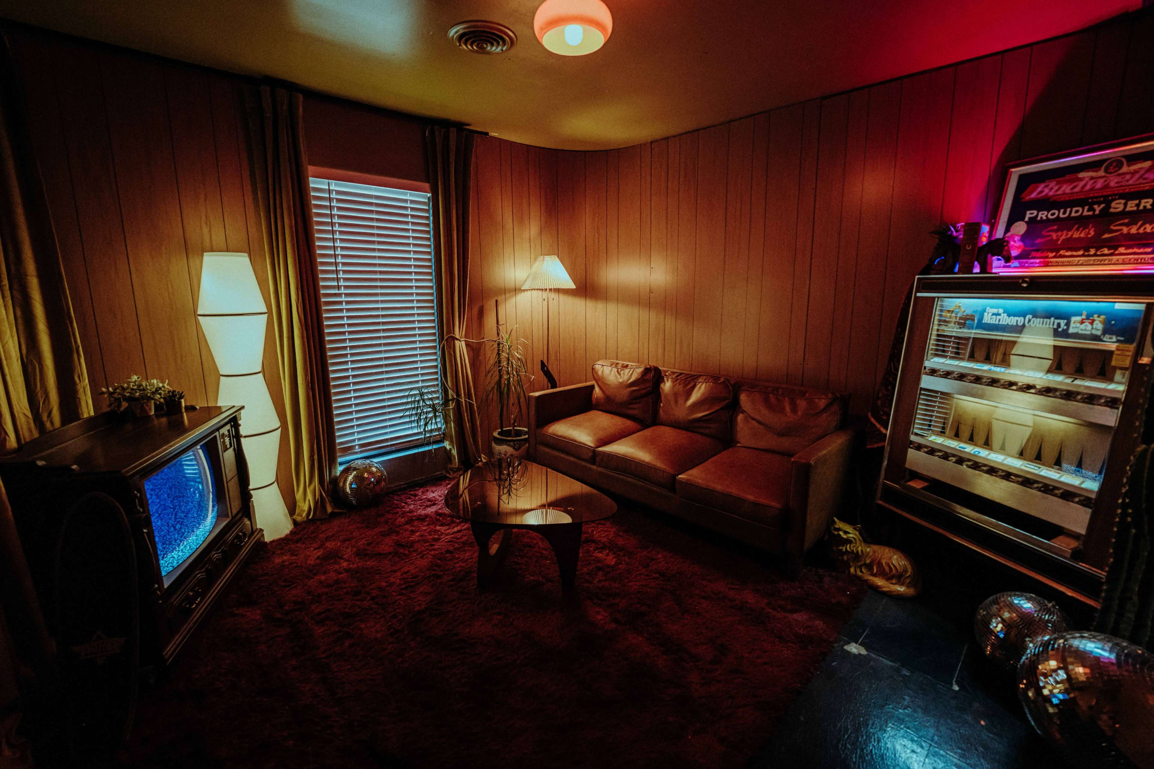 The image shows a retro-style living room featuring a brown leather couch, a round coffee table, a vintage television set, and a small refrigerator beside a window with blinds.