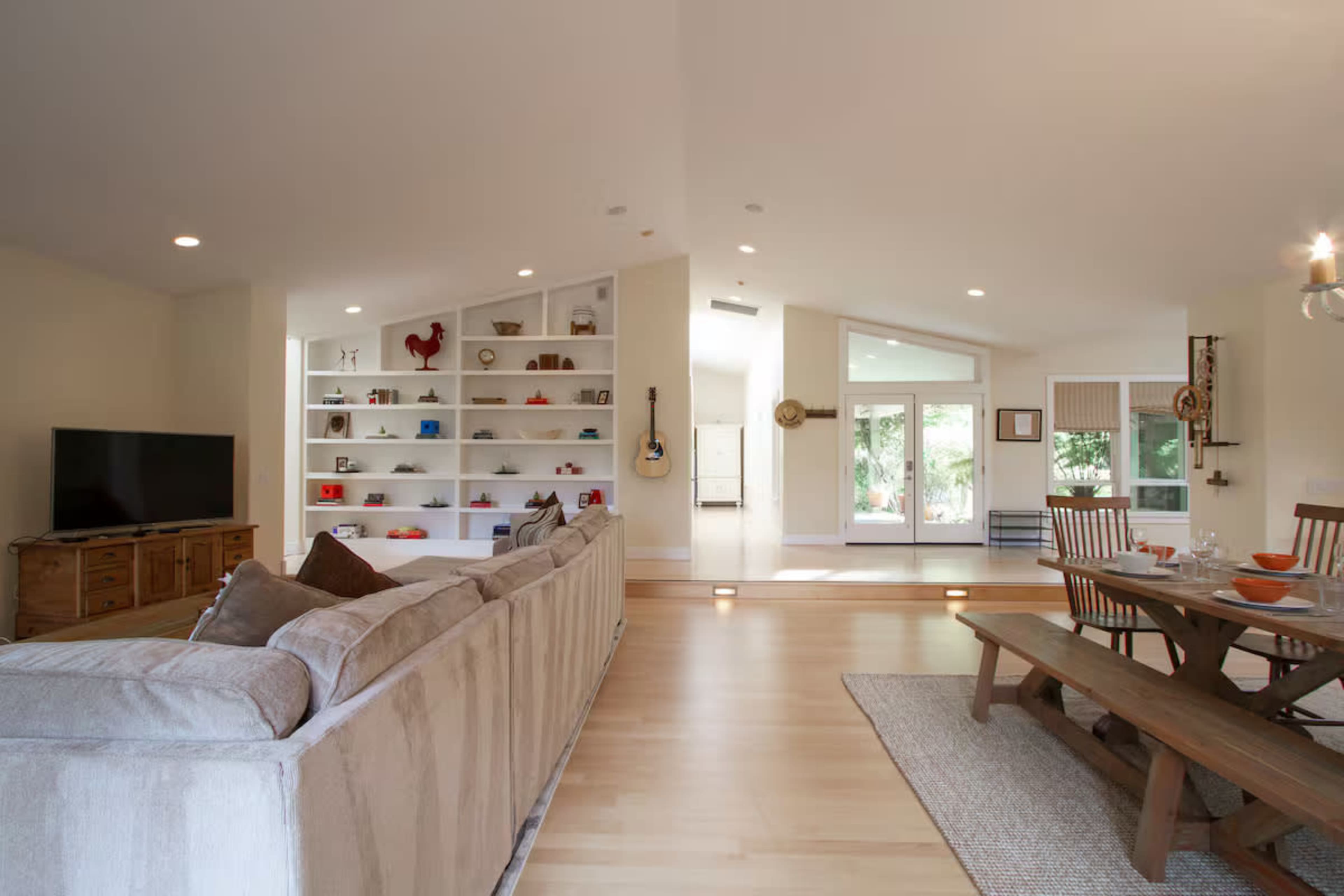 The image features a modern living room and dining area with a beige couch, a wooden dining table, and a shelf displaying decorative items.