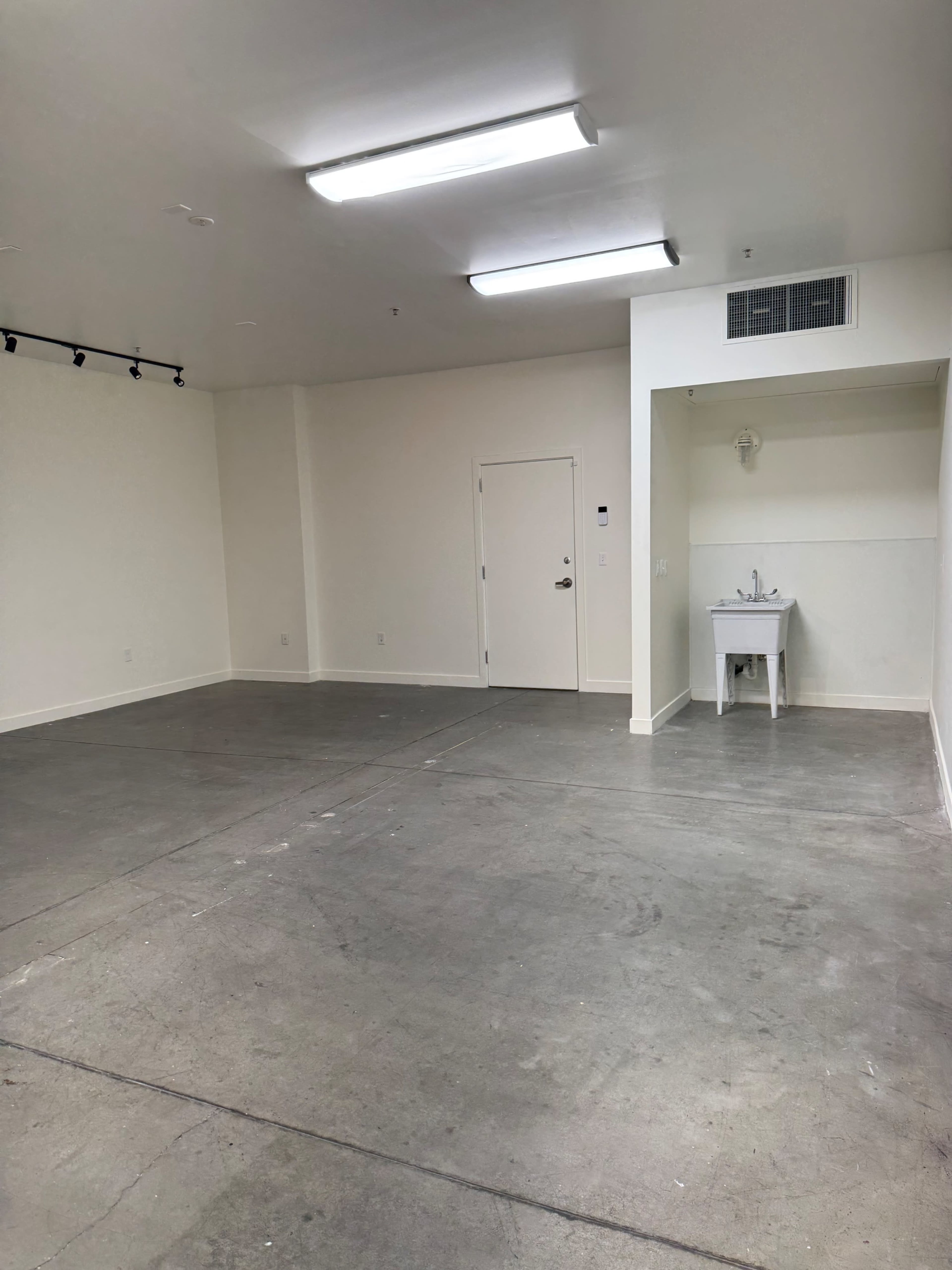 The image shows a spacious, empty room with concrete flooring, white walls, and a small sink area in one corner.