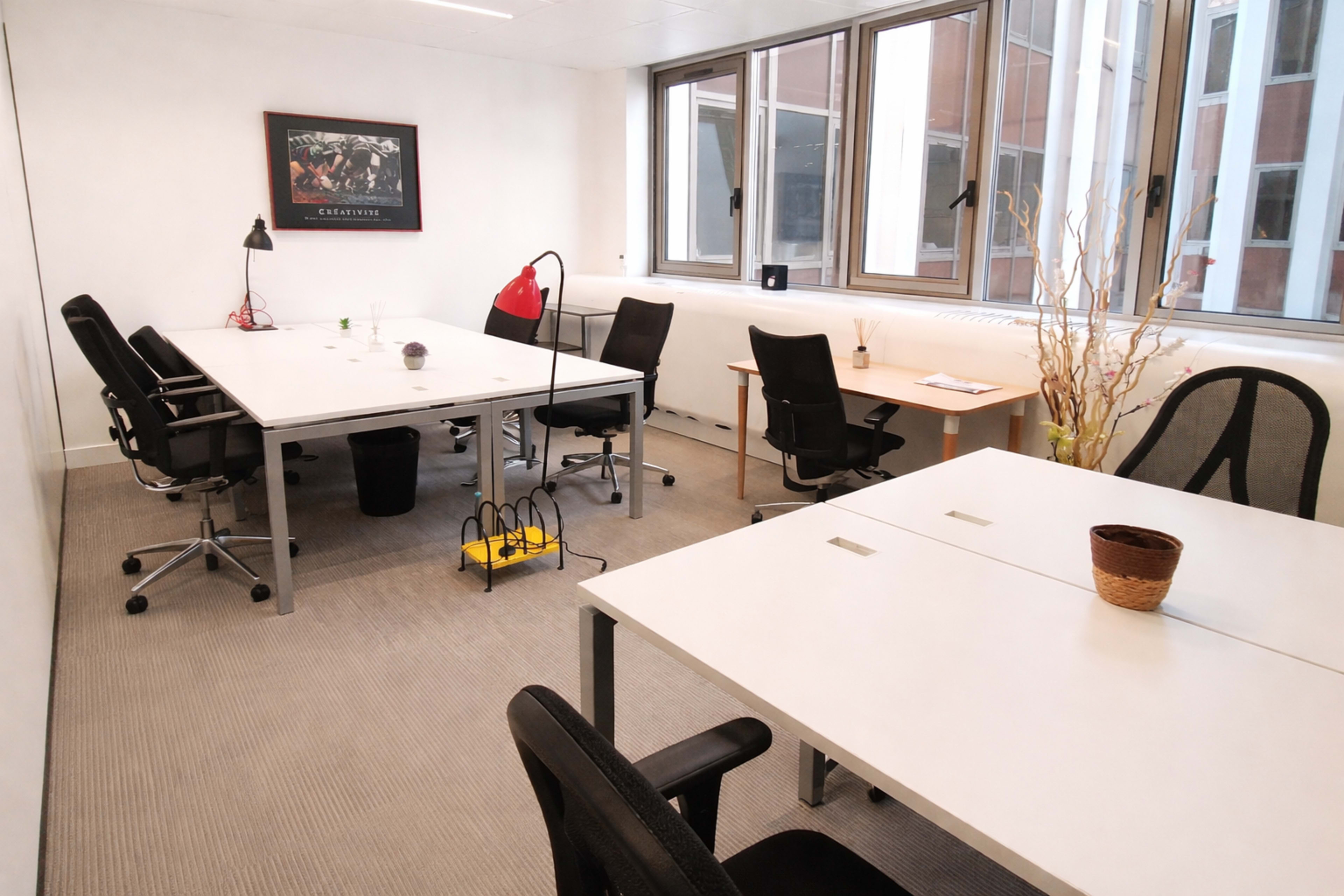 The image shows a modern office space with multiple workstations, including four desks, ergonomic chairs, and large windows that allow natural light to enter.