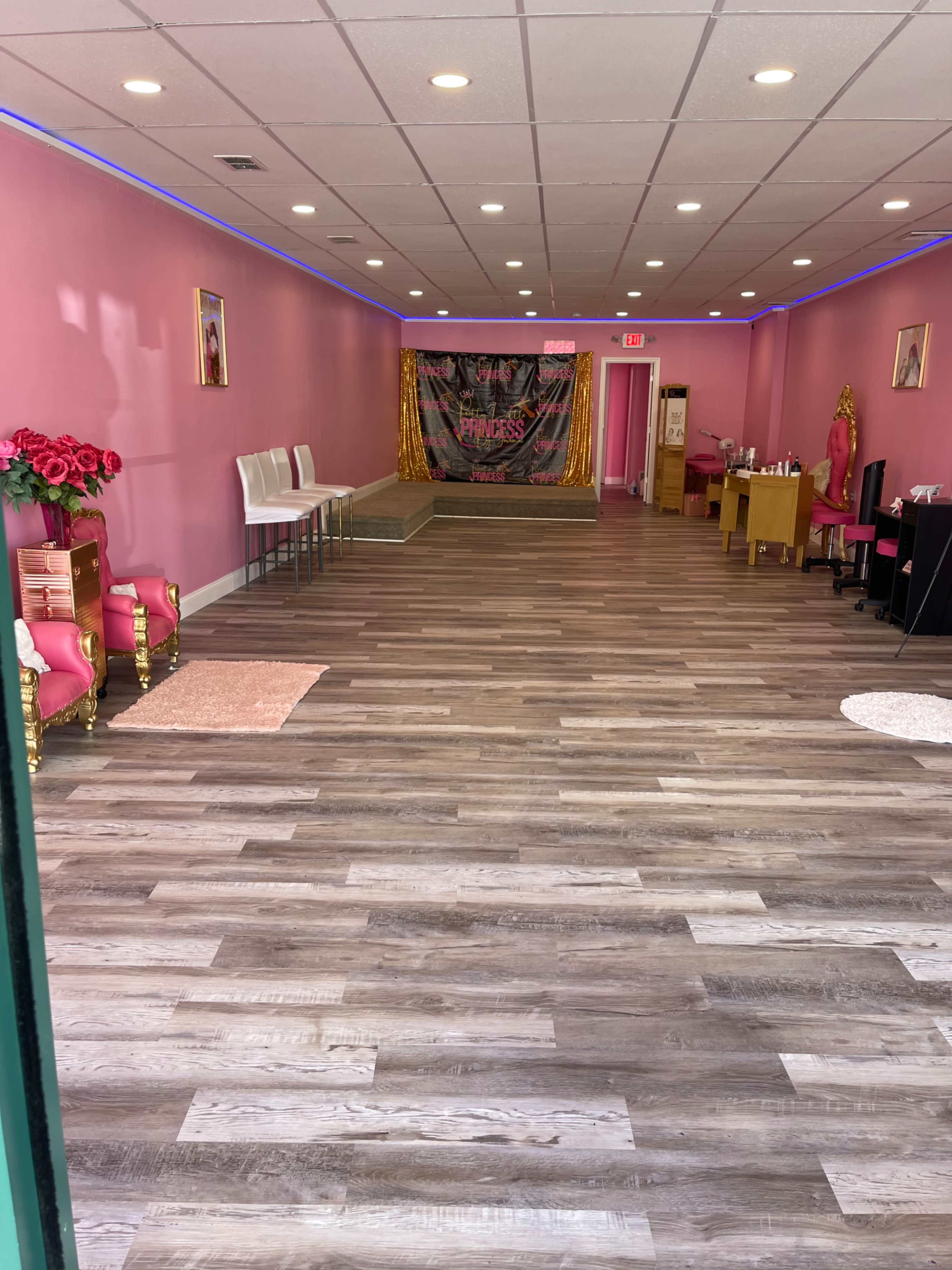 An interior space with pink walls, hardwood flooring, and a backdrop decorated with the word "Princess" at the far end, along with several chairs and a small table on one side.