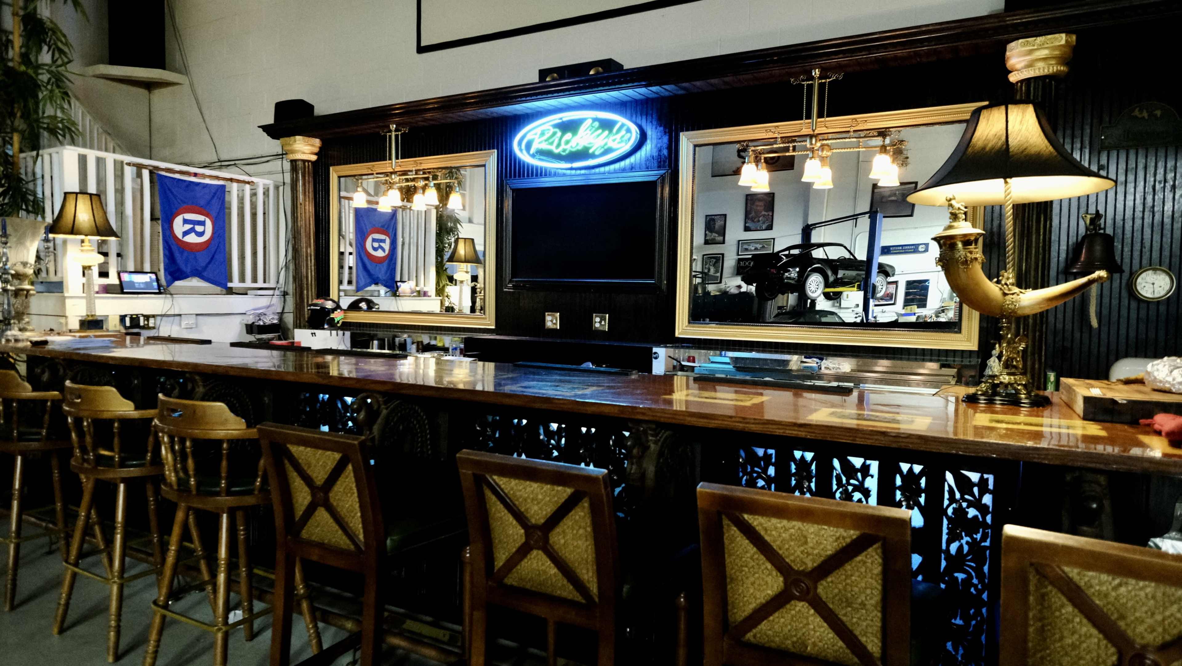 A bar area with a wooden counter, backed by mirrors and illuminated by lamps, featuring baseball-themed decorations.