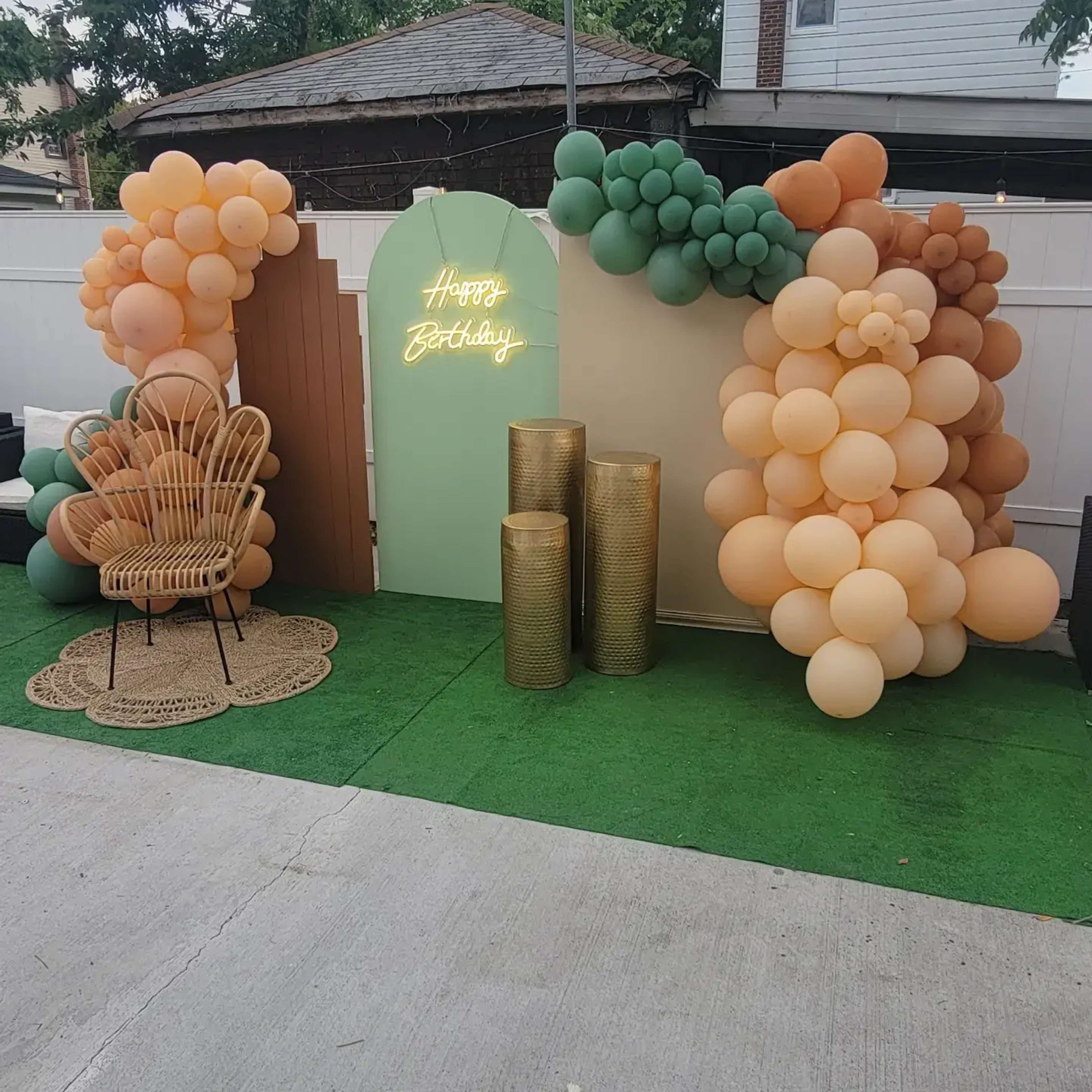 A birthday decoration setup features a backdrop of balloons in various colors, a neon sign proclaiming "Happy Birthday," a rattan chair, and metallic decorative columns on a grassy surface.