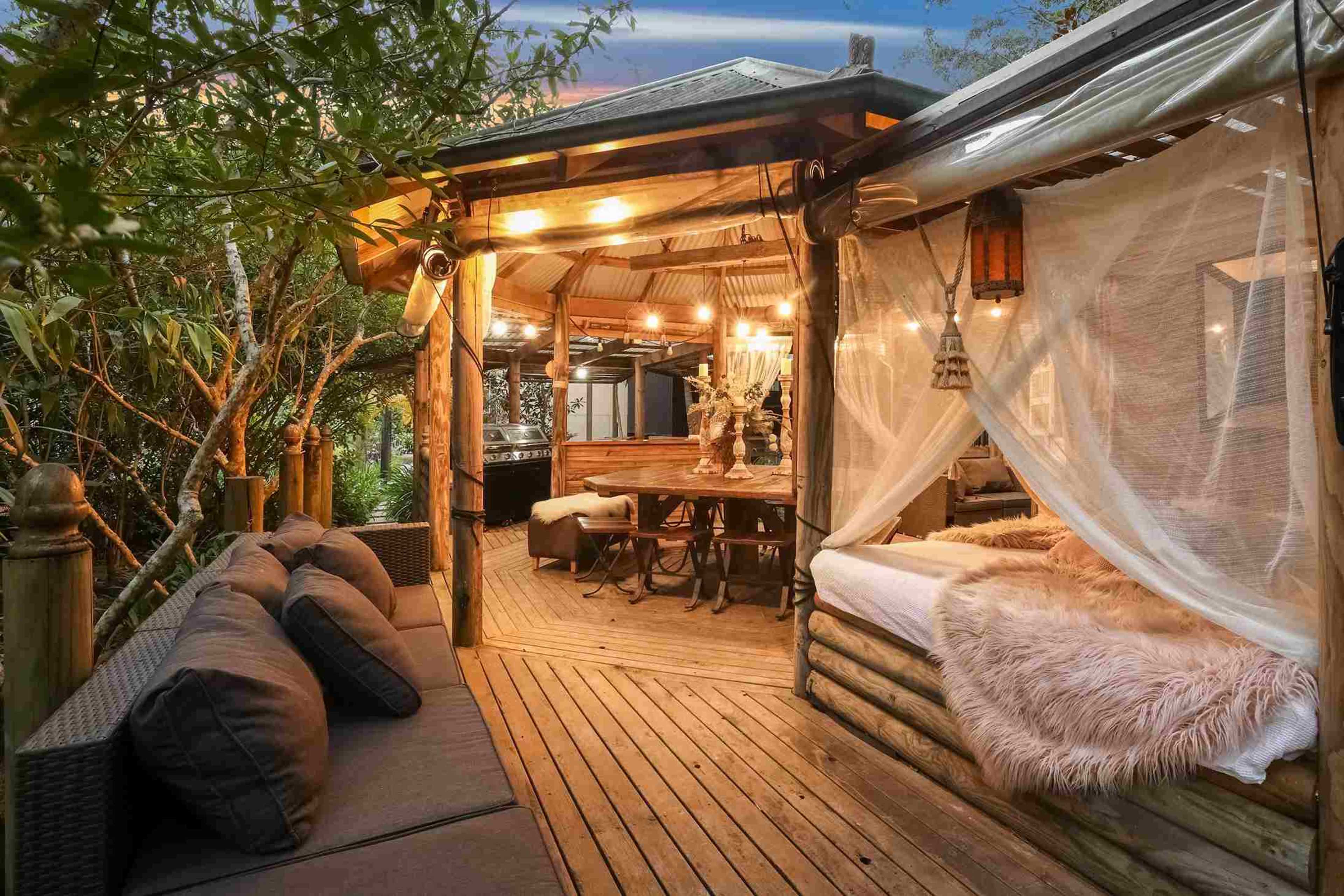A wooden deck with a cozy seating area, a dining space, and a bed framed by sheer curtains, surrounded by greenery and softly glowing lights.