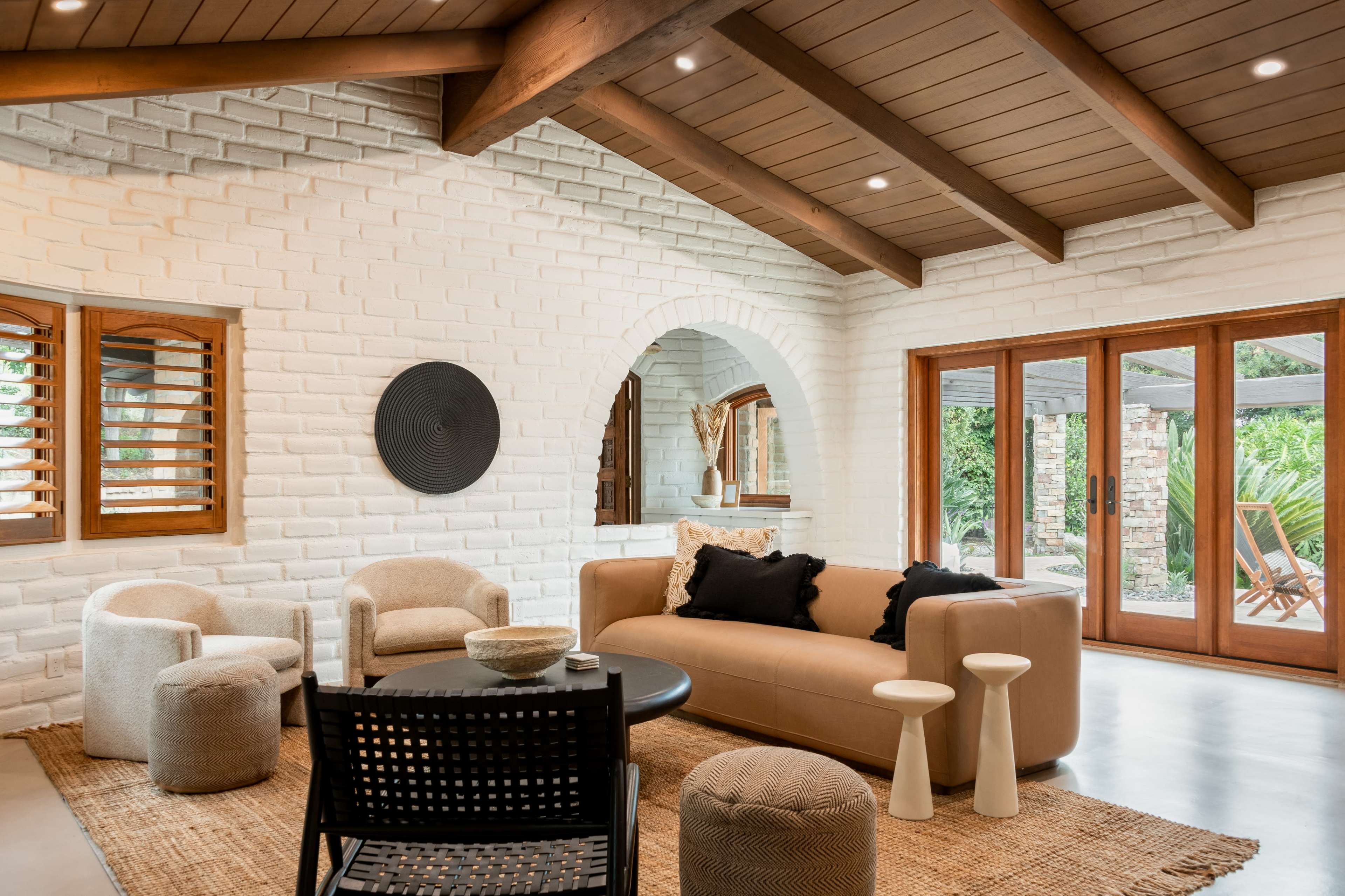 A modern living room with a beige sofa, two small wooden side tables, and several cozy chairs, all arranged around a circular black coffee table, with glass doors leading to an outdoor area.
