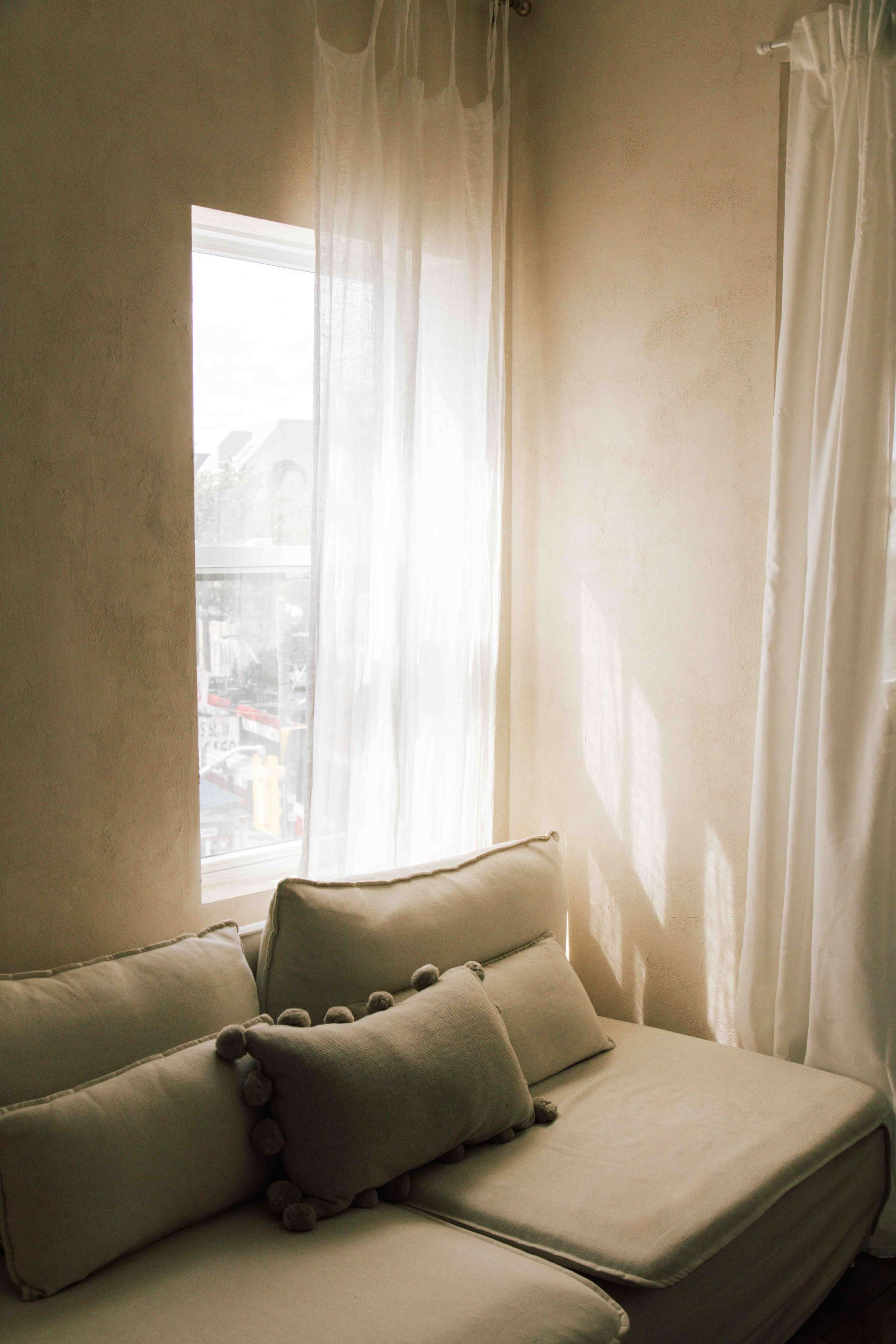A sunlit corner of a room features a sofa with decorative pillows and sheer curtains by a window.