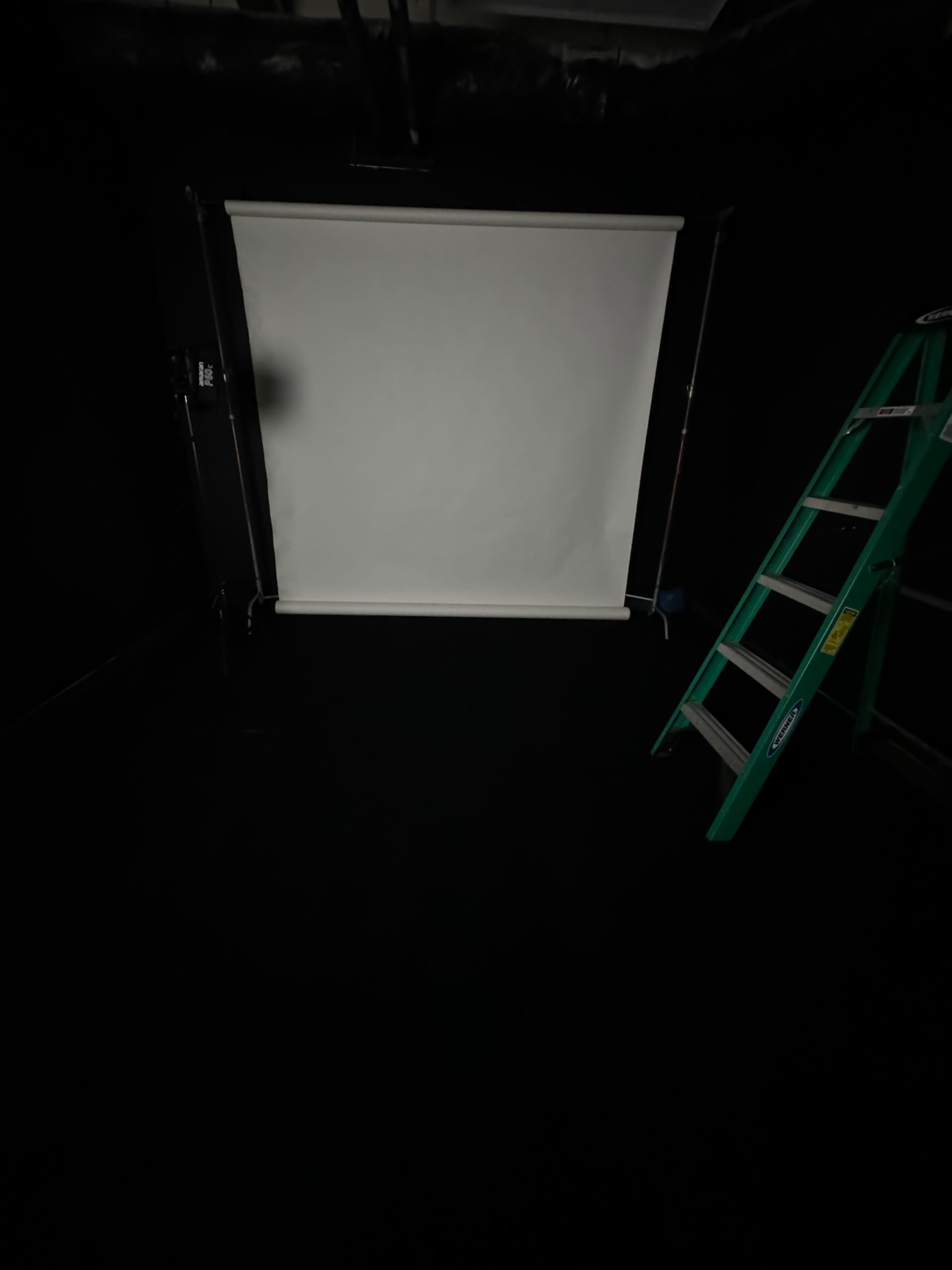 The image shows a dark room with a white backdrop on a stand and a green ladder positioned nearby.