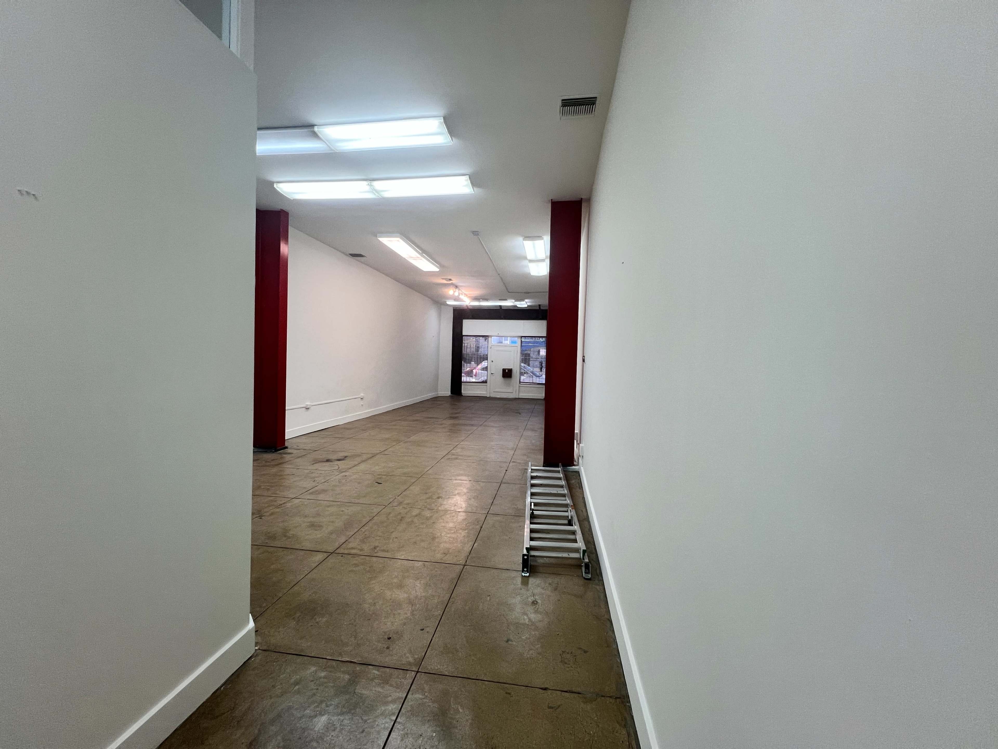 The image shows a long, empty hallway leading to a large open space with a screen at the end, illuminated by overhead lights.