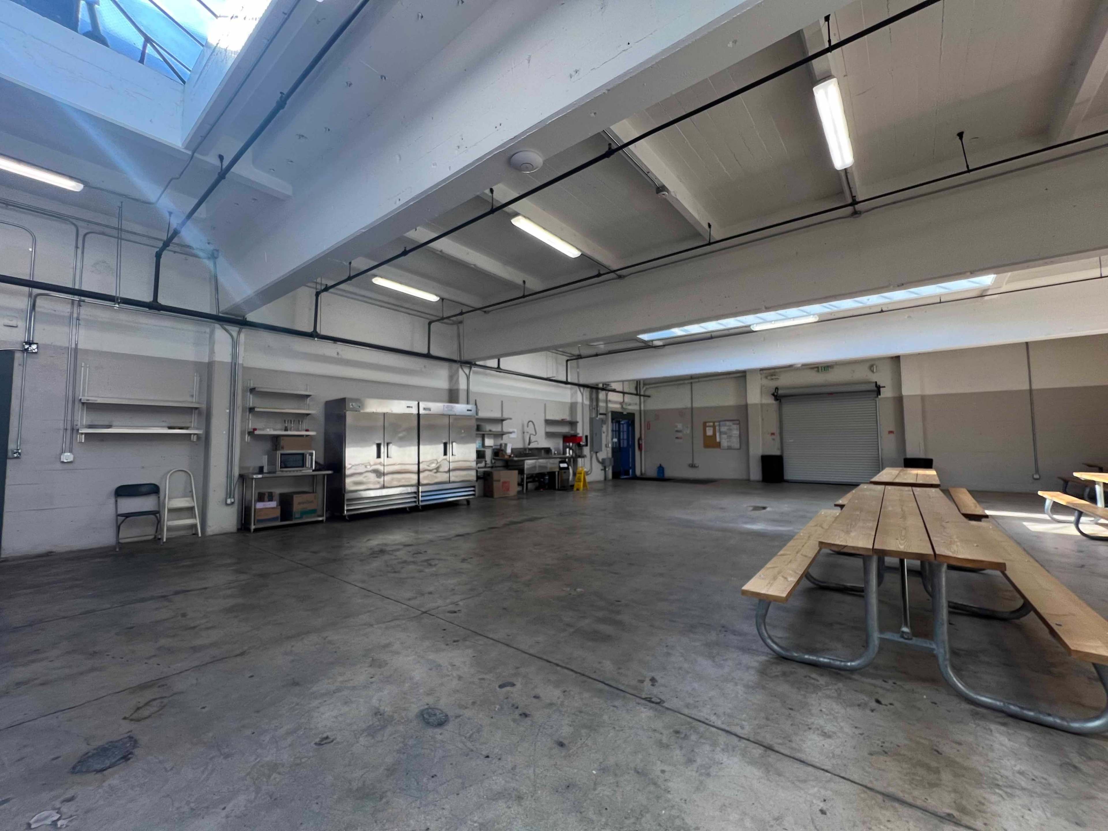 An empty industrial kitchen area with stainless steel appliances and wooden picnic-style tables.