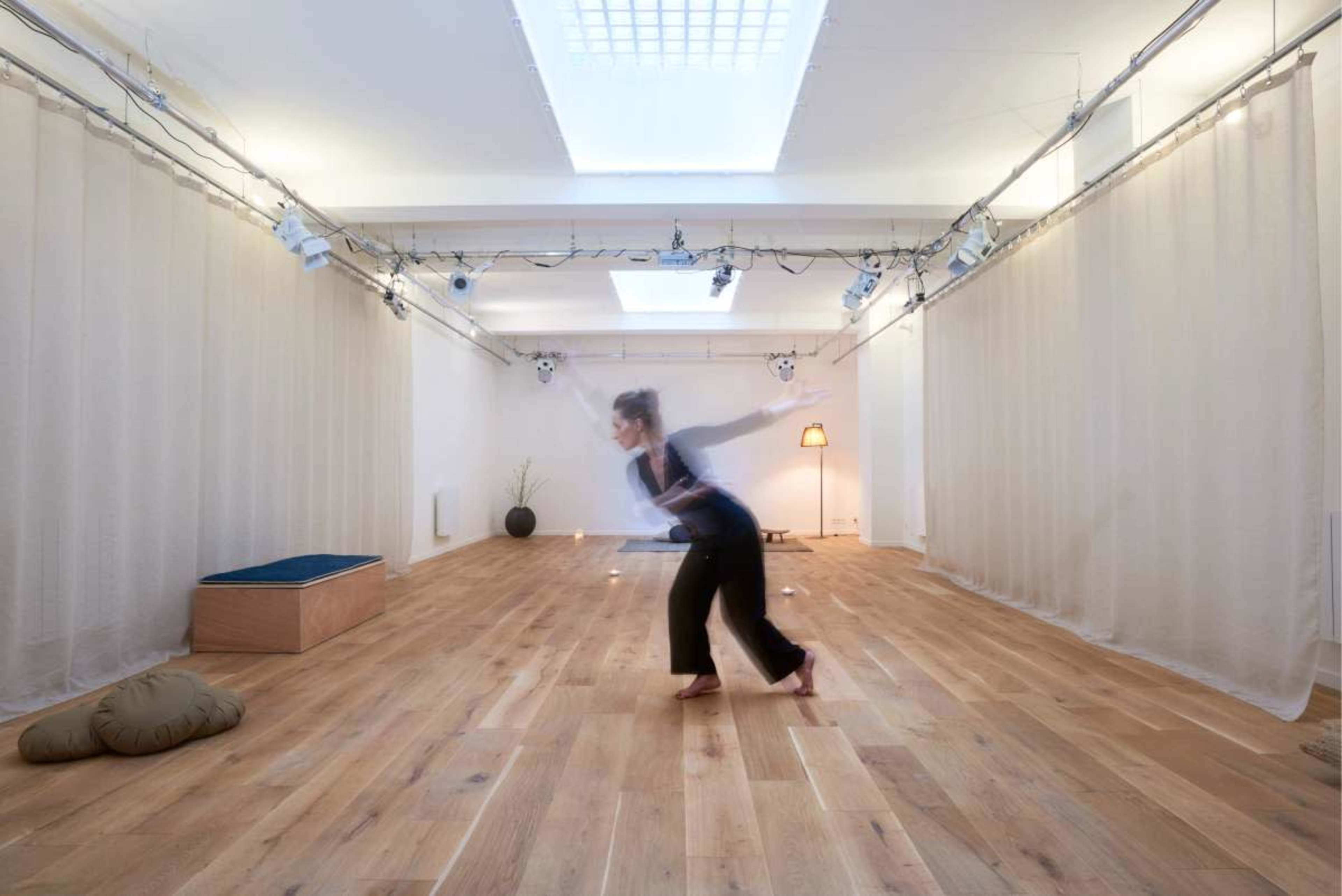 A person is dancing in a spacious, well-lit studio with wooden floors and sheer curtains.