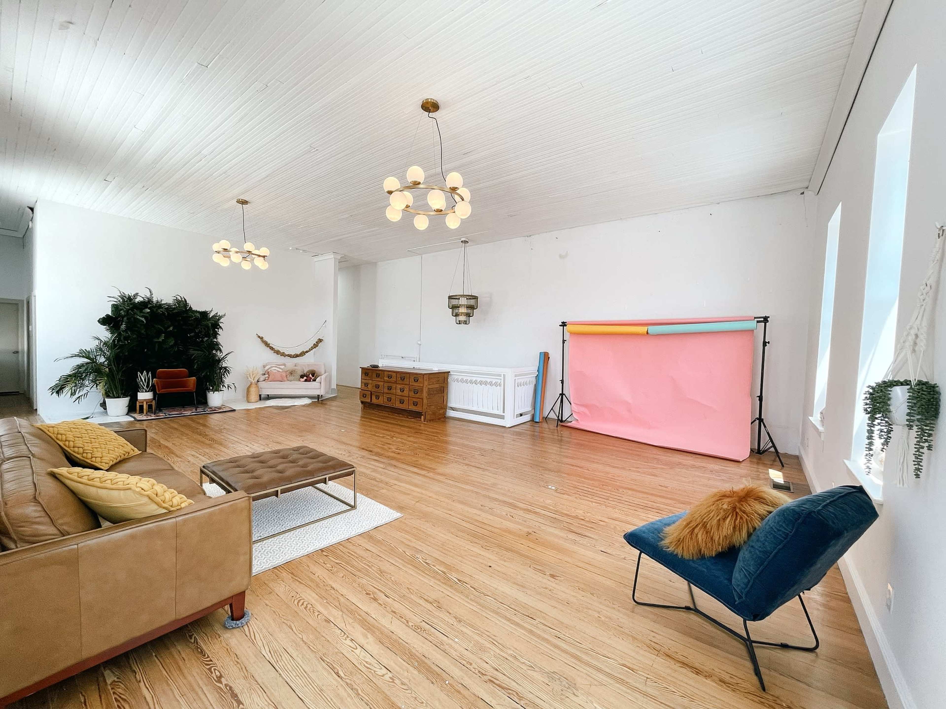 A spacious, well-lit room features wooden floors, a large sofa, and a pink backdrop with lighting equipment set up against a white wall.