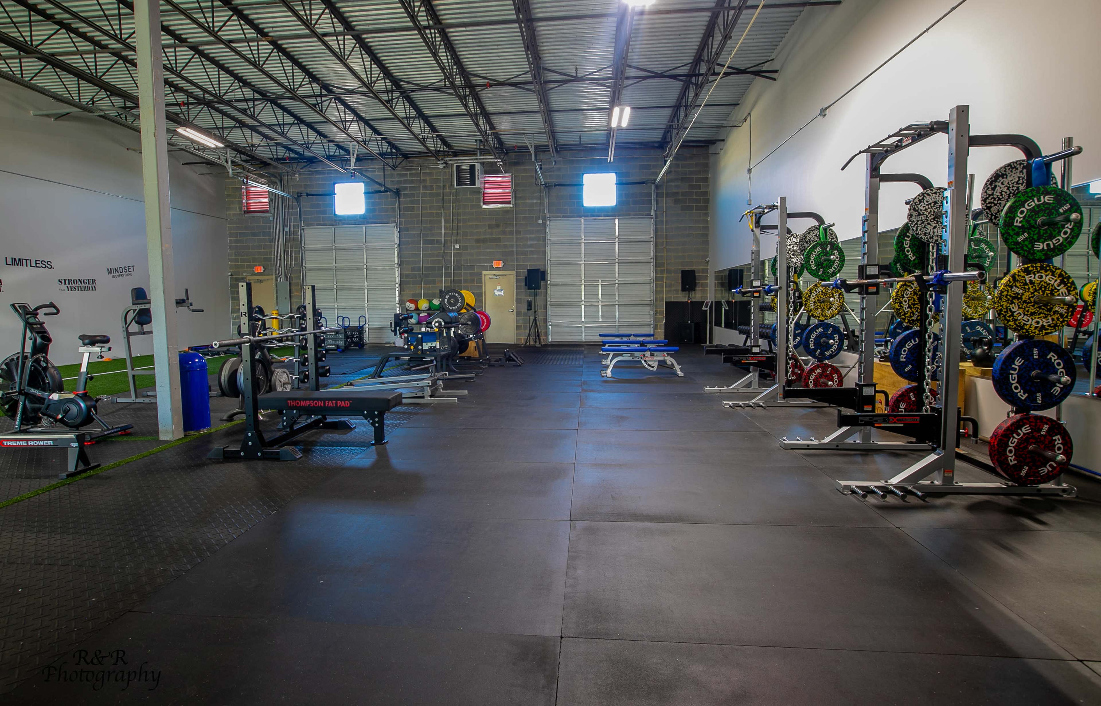 The image shows a spacious indoor gym with various weightlifting equipment, benches, and exercise mats arranged neatly along the walls.
