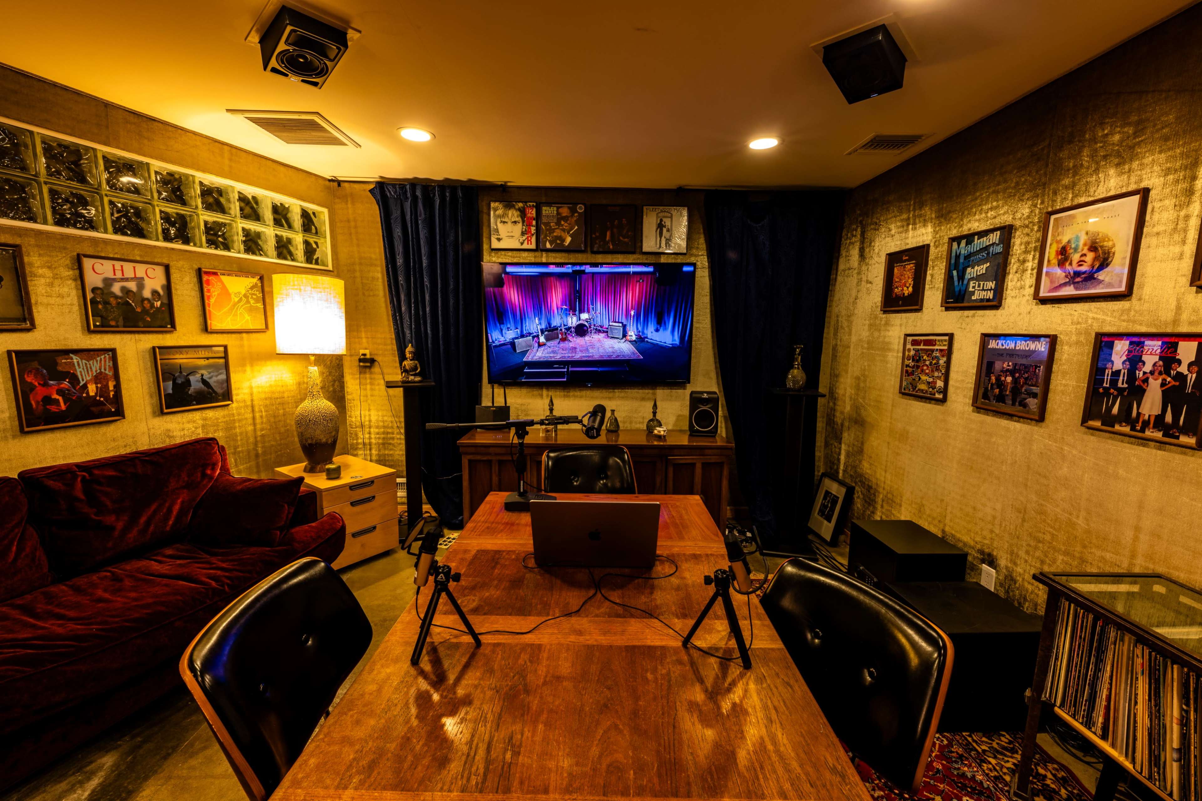 The image shows a small recording studio or podcast room with a wooden table, black chairs, a large screen displaying a performance, and various framed records on the walls.