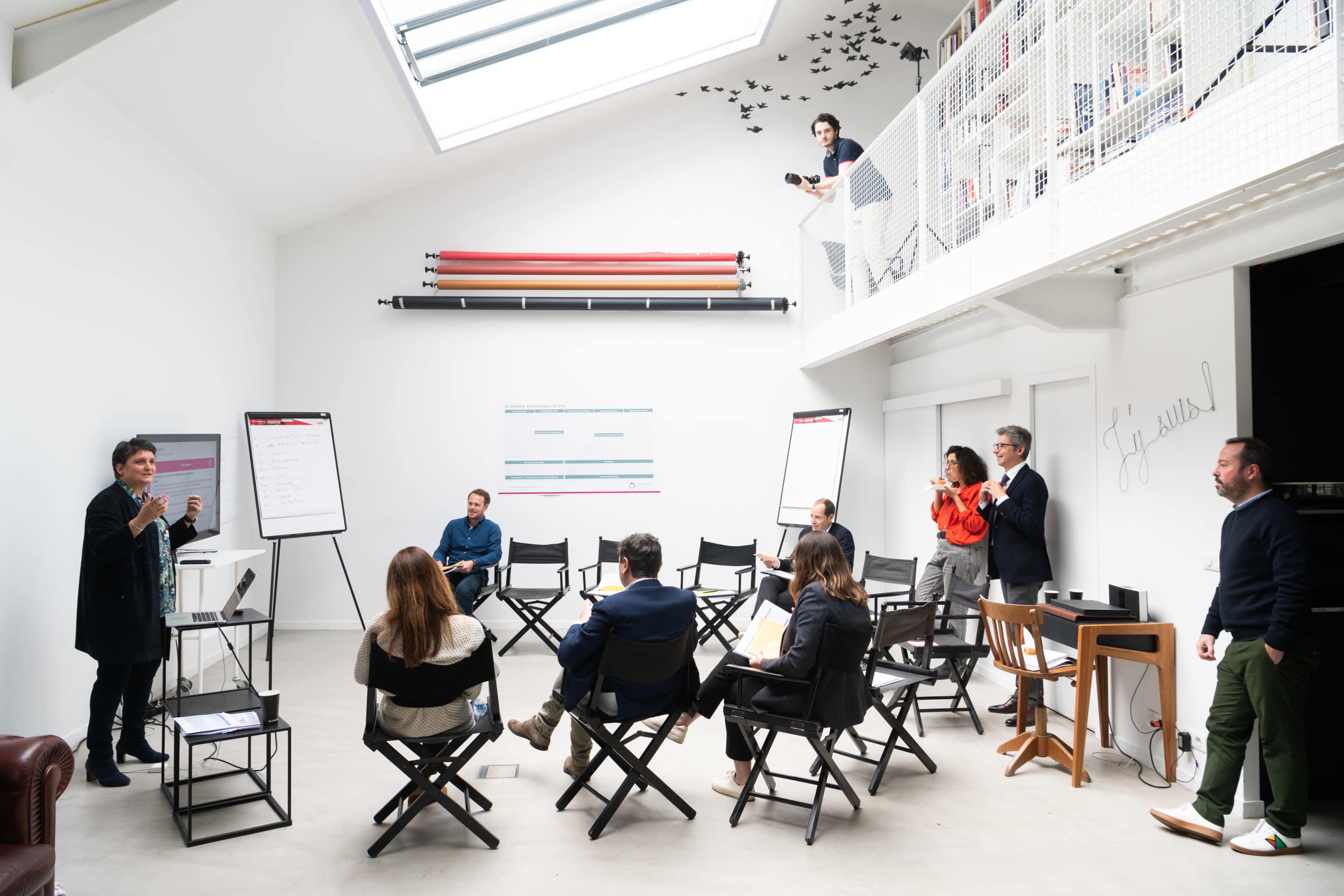 A group of people engage in a meeting in a bright, modern room with a skylight and a whiteboard.