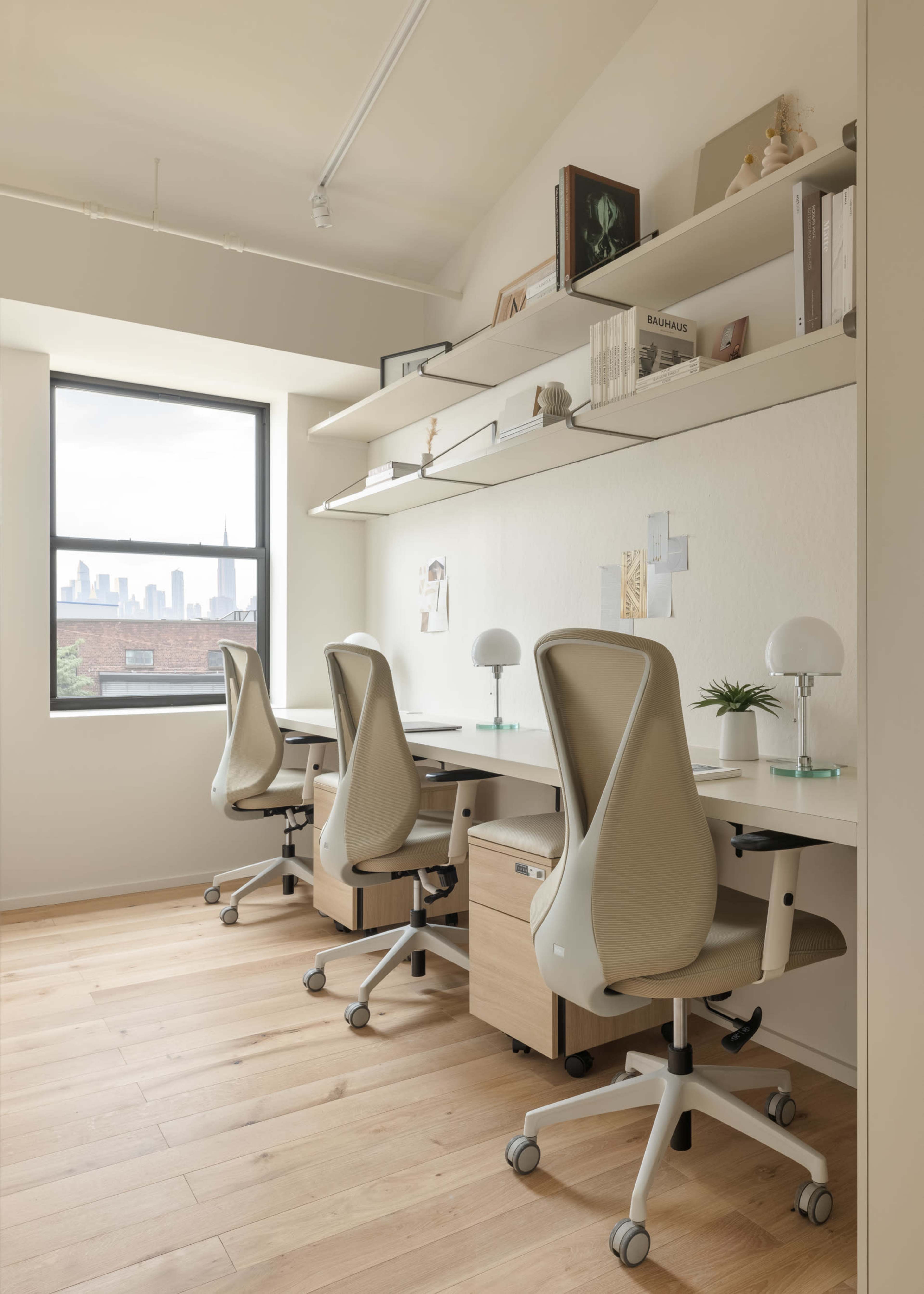 The image shows a modern office space with three ergonomic chairs at individual desks, a window with a city skyline view, and shelves neatly arranged above the desks.