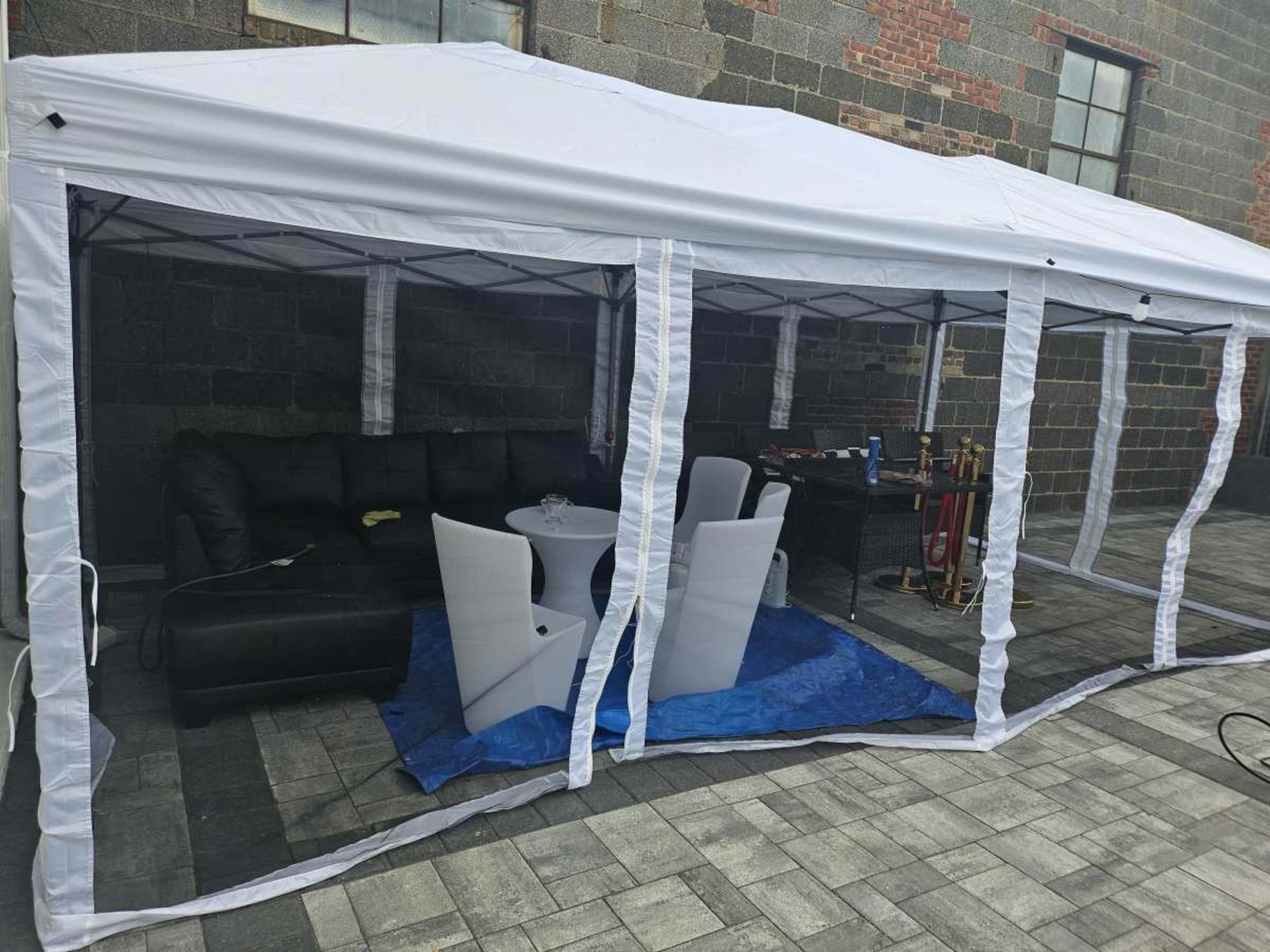 A white canopy tent is set up outside, featuring a black couch, white chairs, and a table on a paved surface.