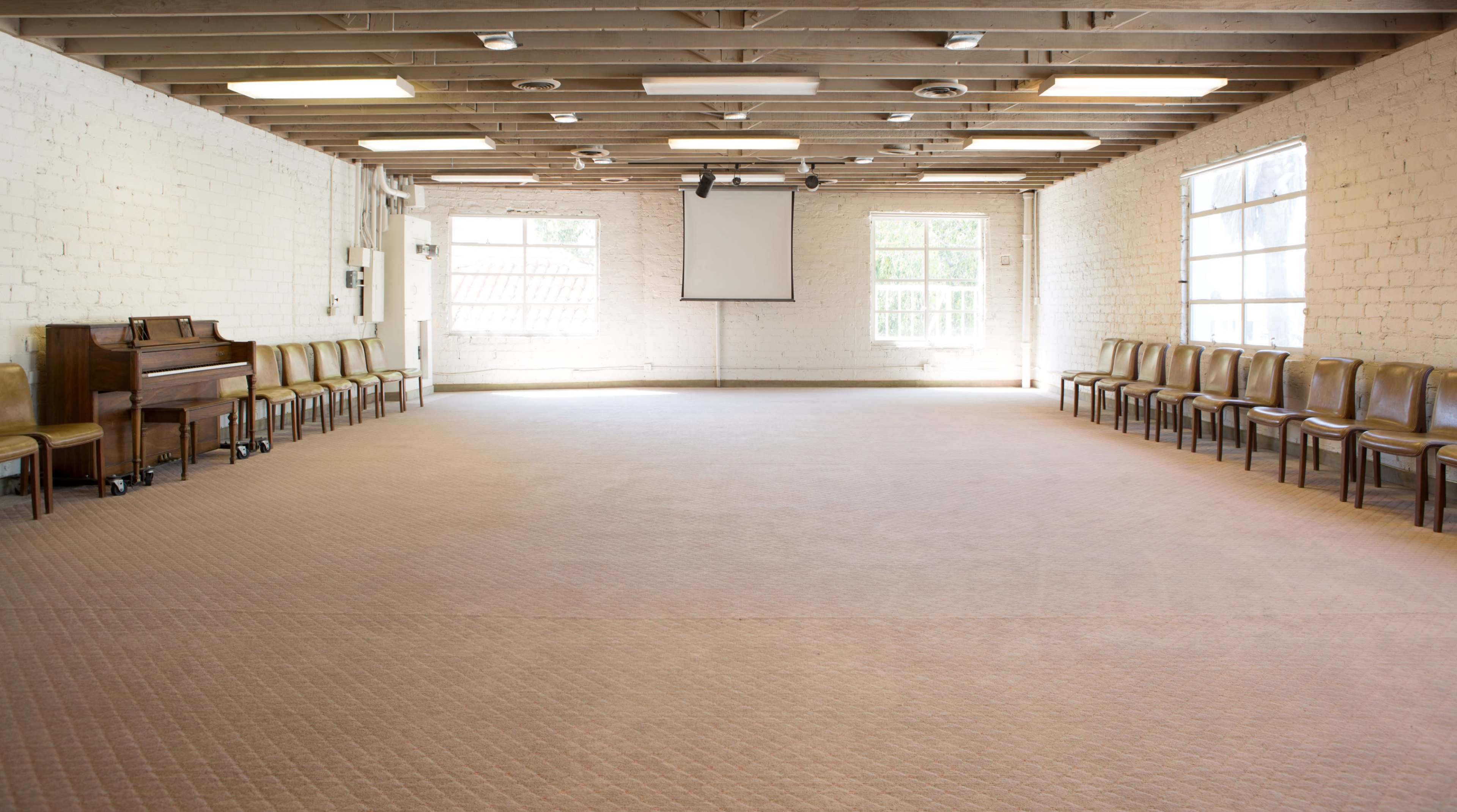 A spacious room with a polished floor, a row of chairs along one side, and a piano positioned nearby under a projection screen.