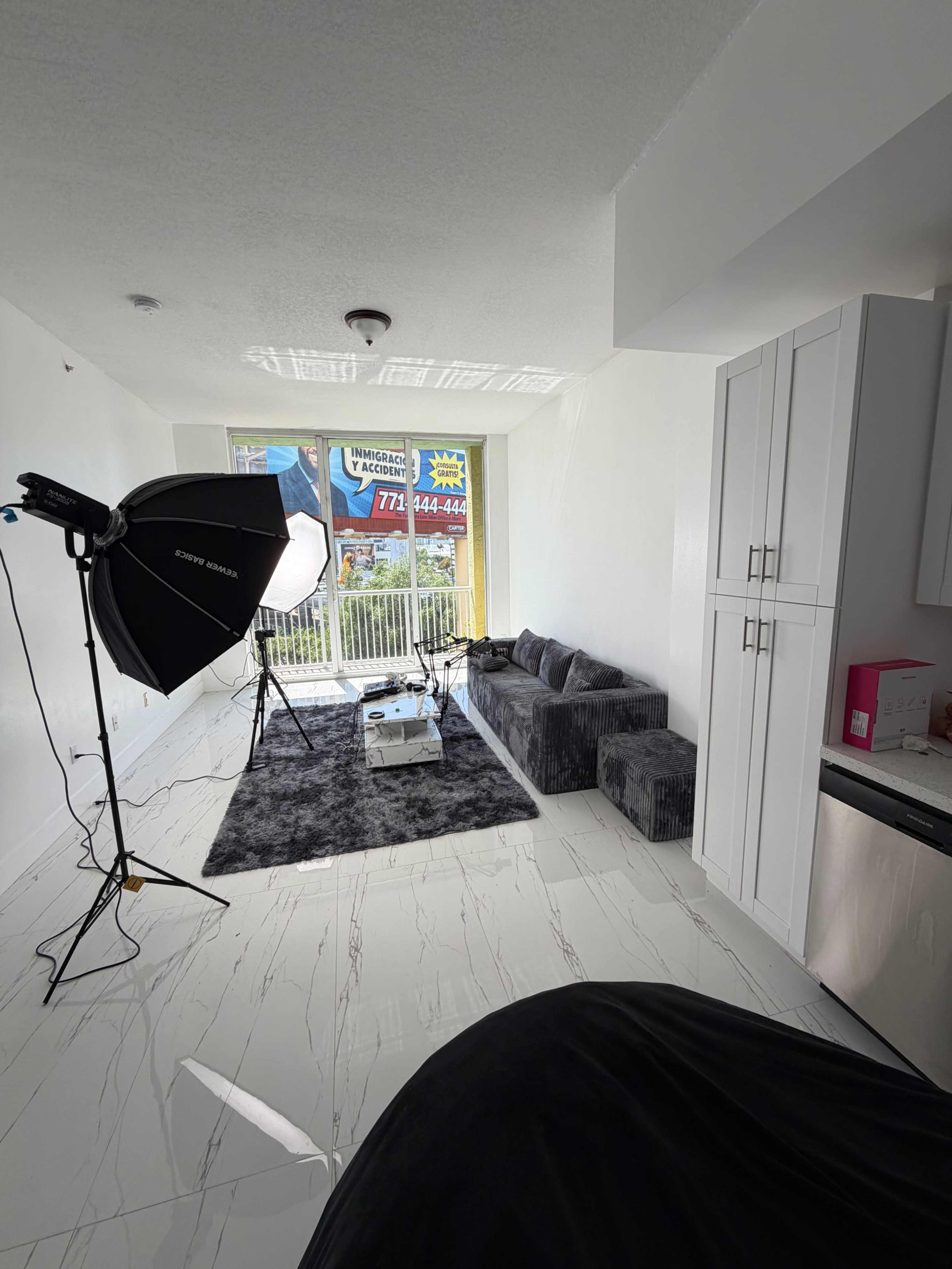 A well-lit room features a gray sofa, a rug, and a lighting setup in front of a large window with visible signs.