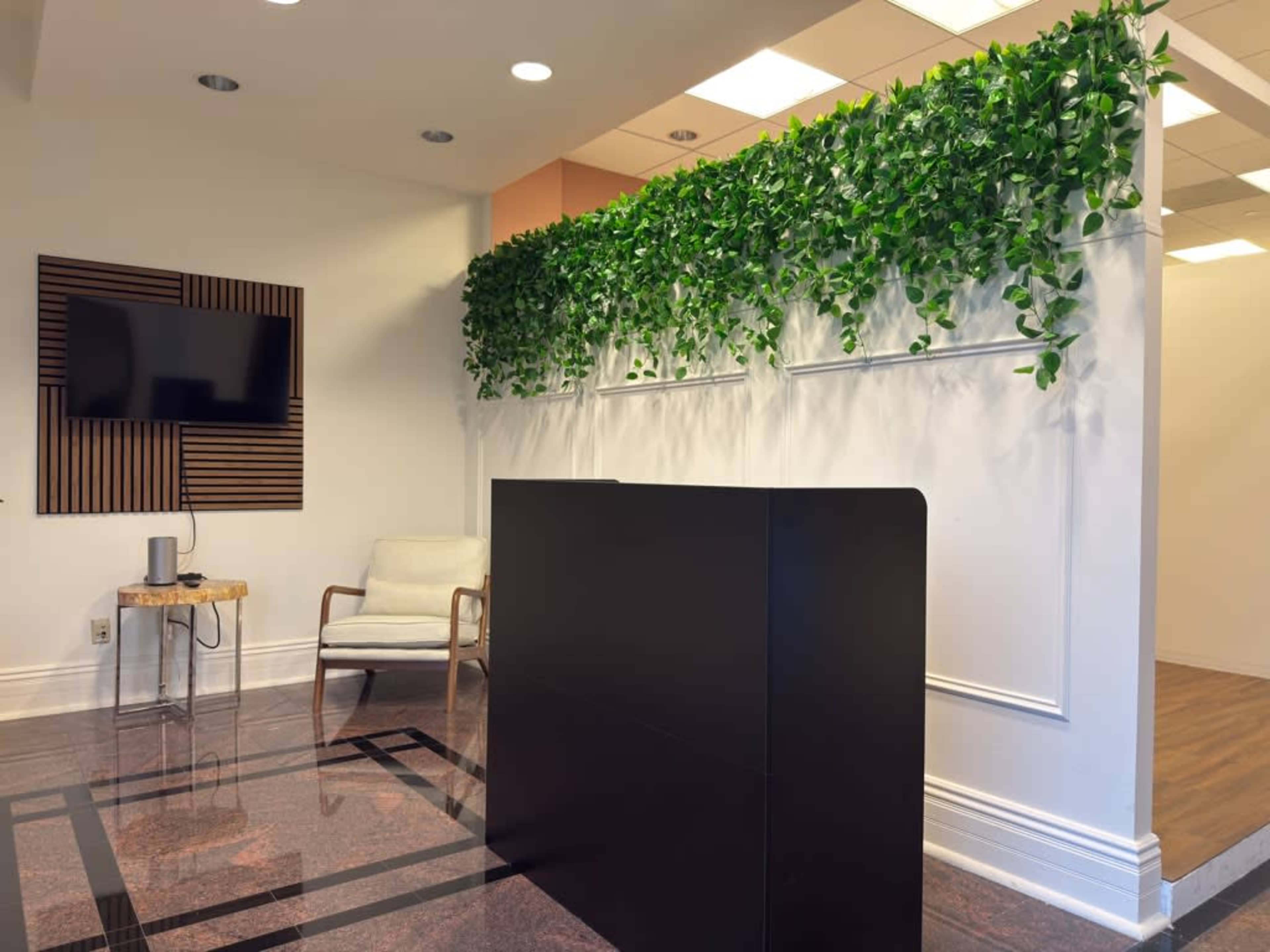 The image shows a modern reception area with a black front desk, a green plant wall, and a television mounted on the wall.