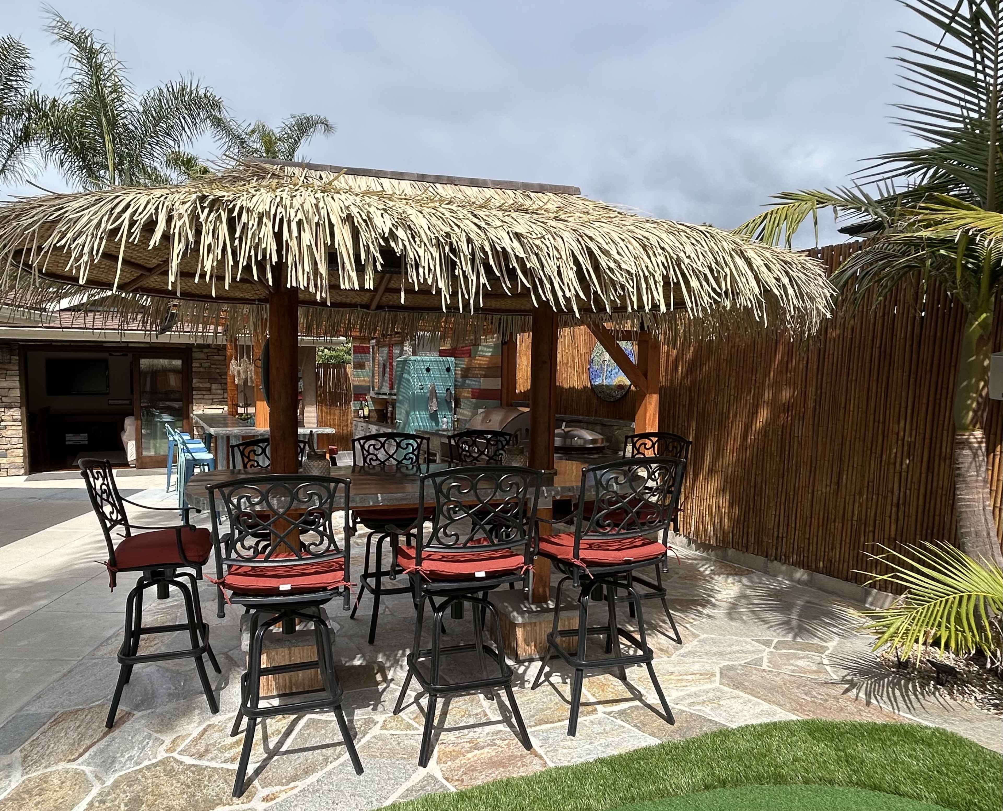 A thatched-roof gazebo with a wooden table and chairs is surrounded by tropical landscaping and stone flooring.