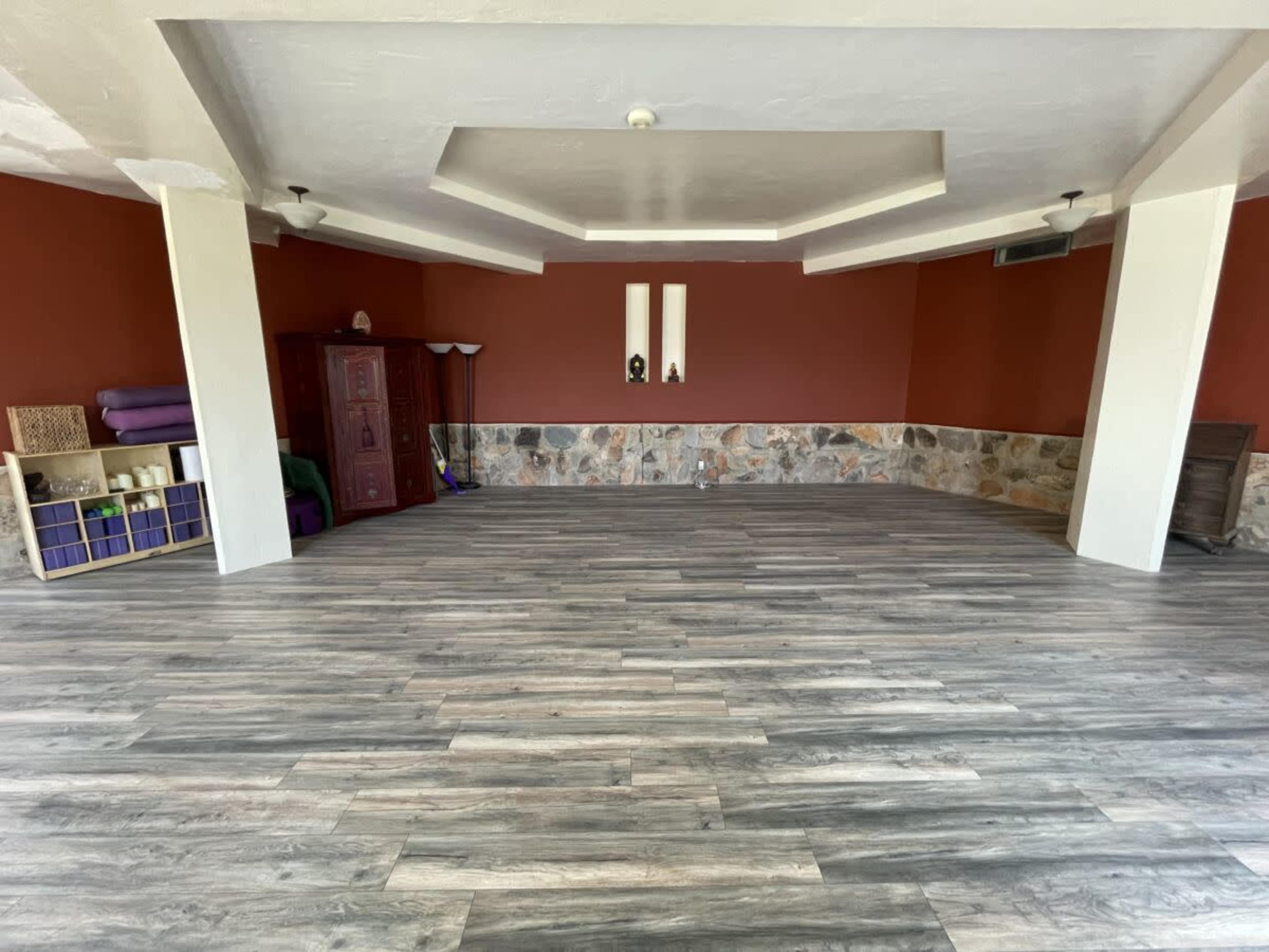The image shows a spacious interior room with wooden flooring, orange walls, and a stone accent wall, featuring yoga mats and storage furniture in the corners.