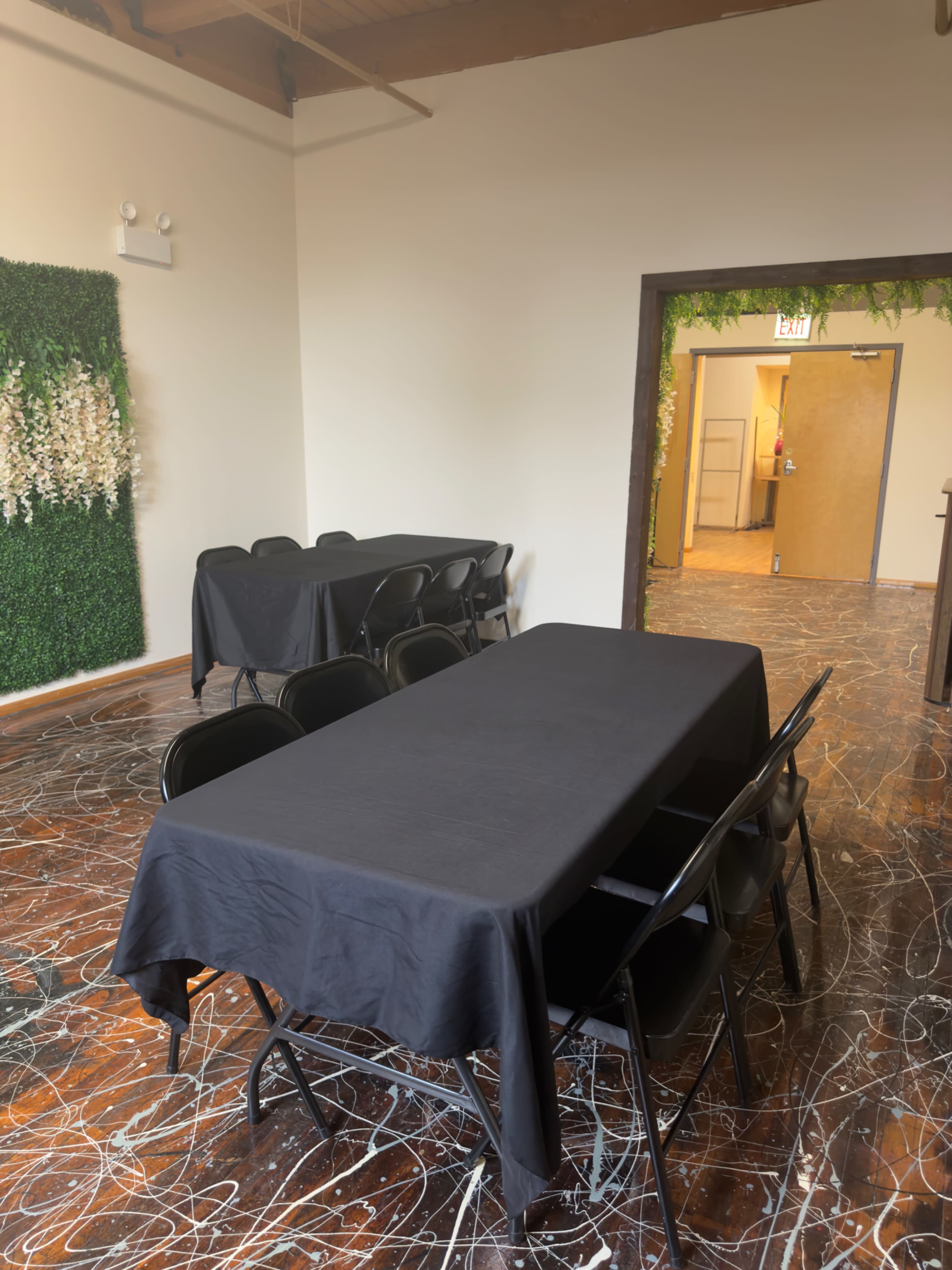 The image shows an empty room with several black-covered tables and chairs, along with a door leading to another area in the background.