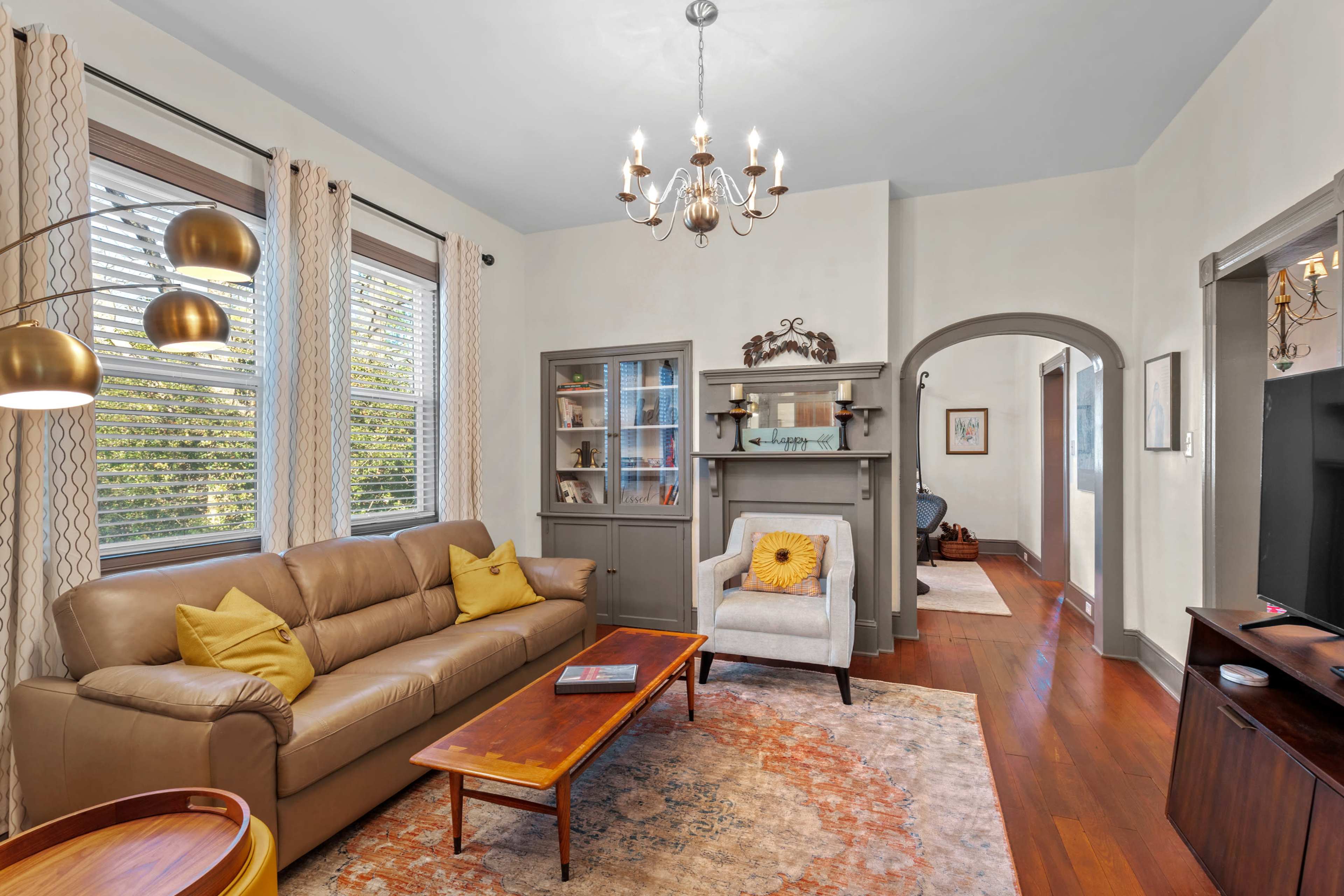 The living room features a beige leather sofa, a wooden coffee table, and a decorative fireplace, with natural light coming through the windows.