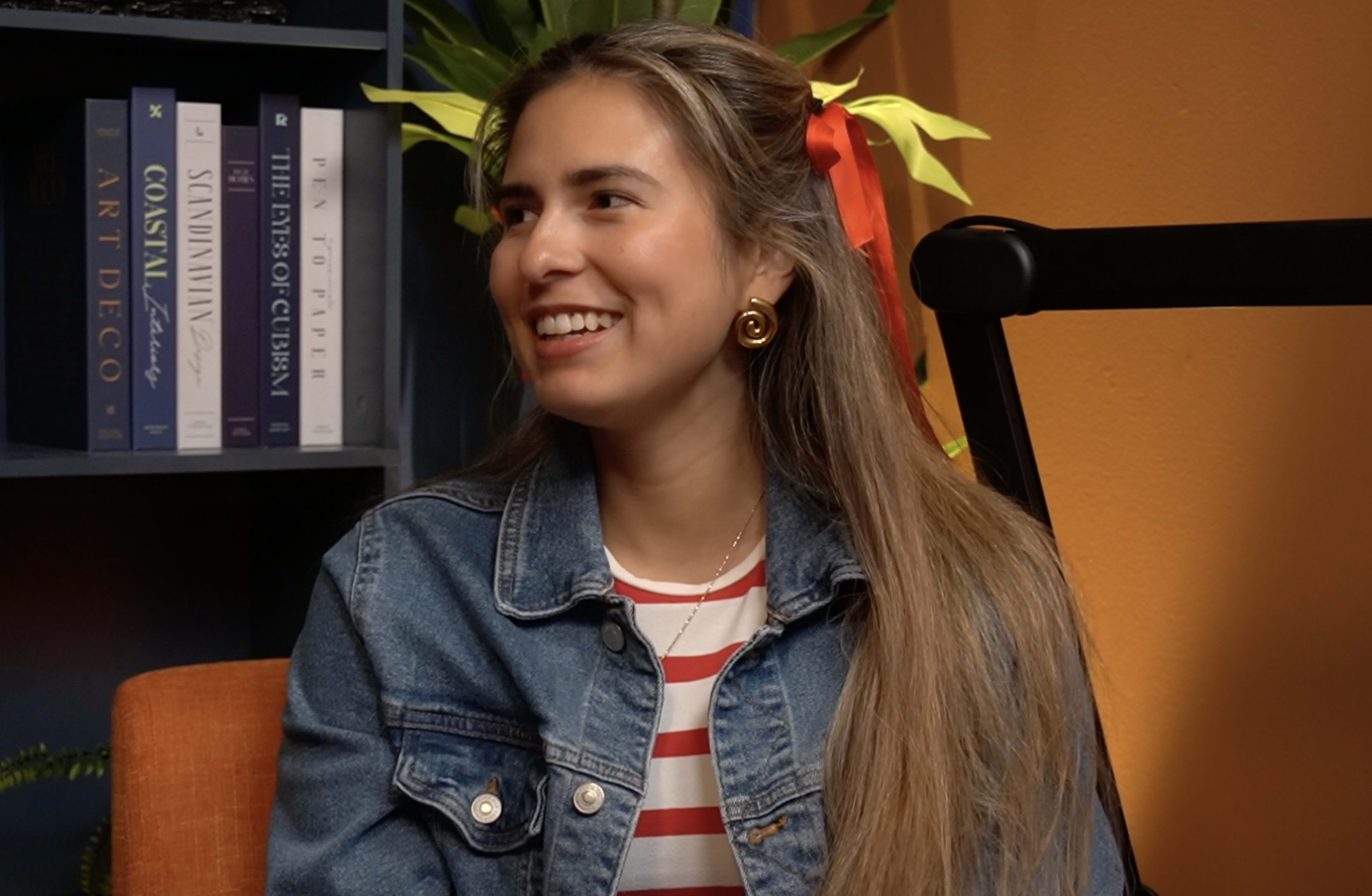 A woman with long hair styled with a ribbon is smiling while wearing a denim jacket over a striped top, seated in front of bookshelves and indoor plants.