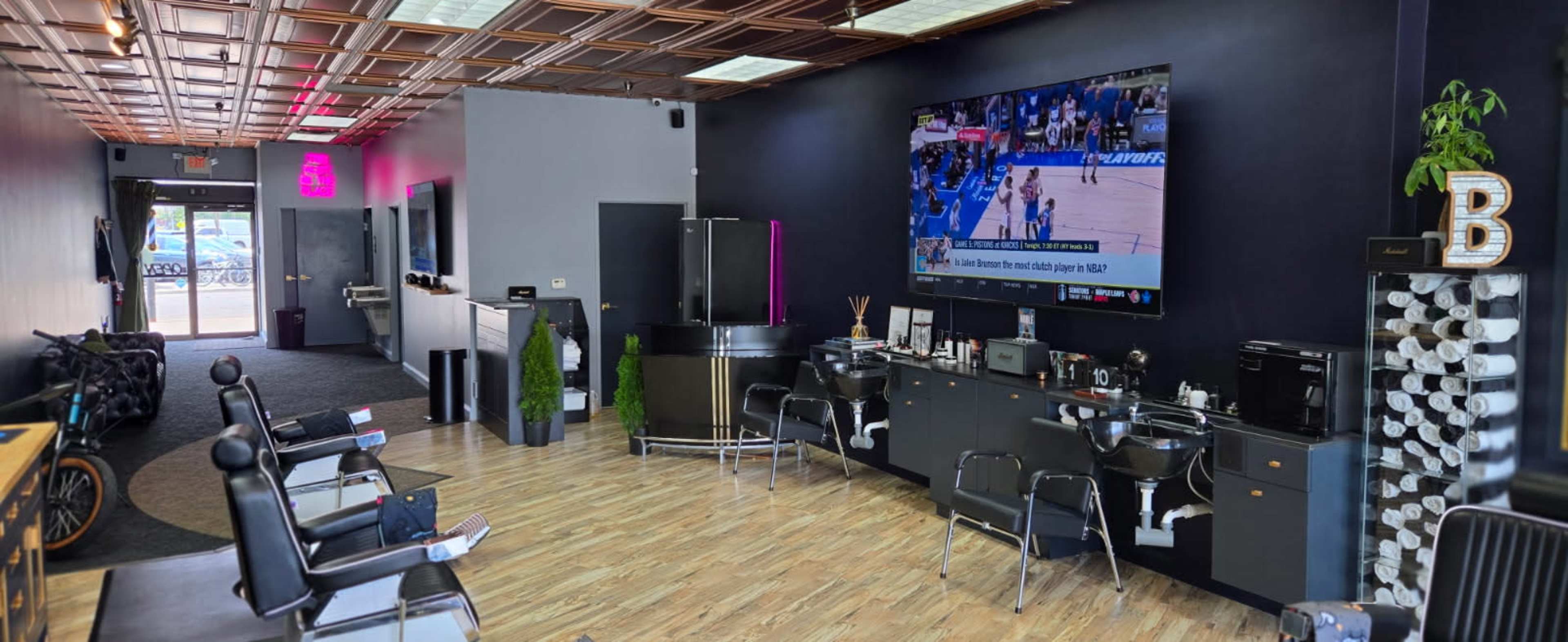 The image shows a modern barbershop interior with black chairs, a large television displaying a sports event, and a sleek reception area.