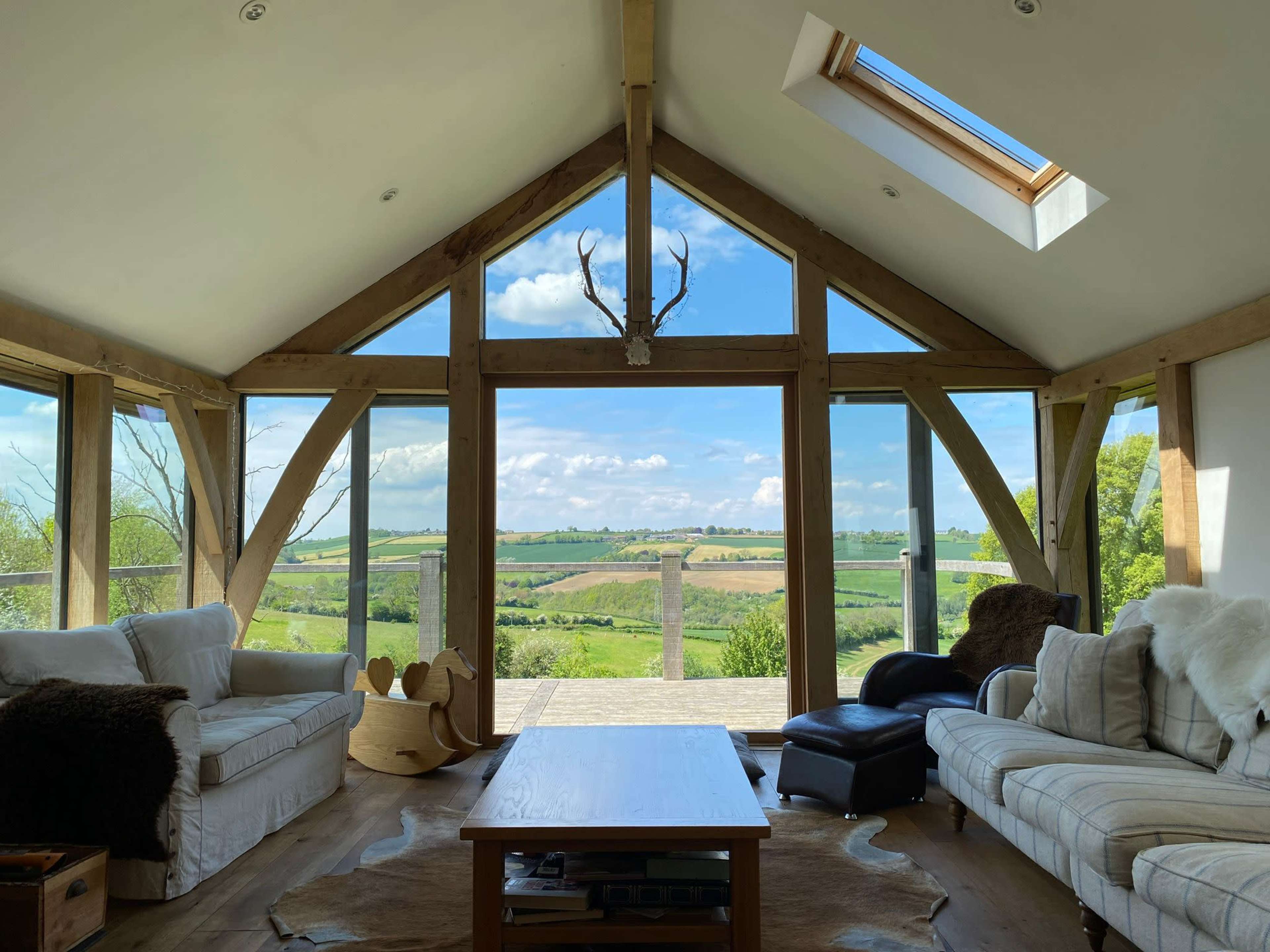 The image shows a modern living room with large windows overlooking a green landscape and wooden beams supporting the ceiling.
