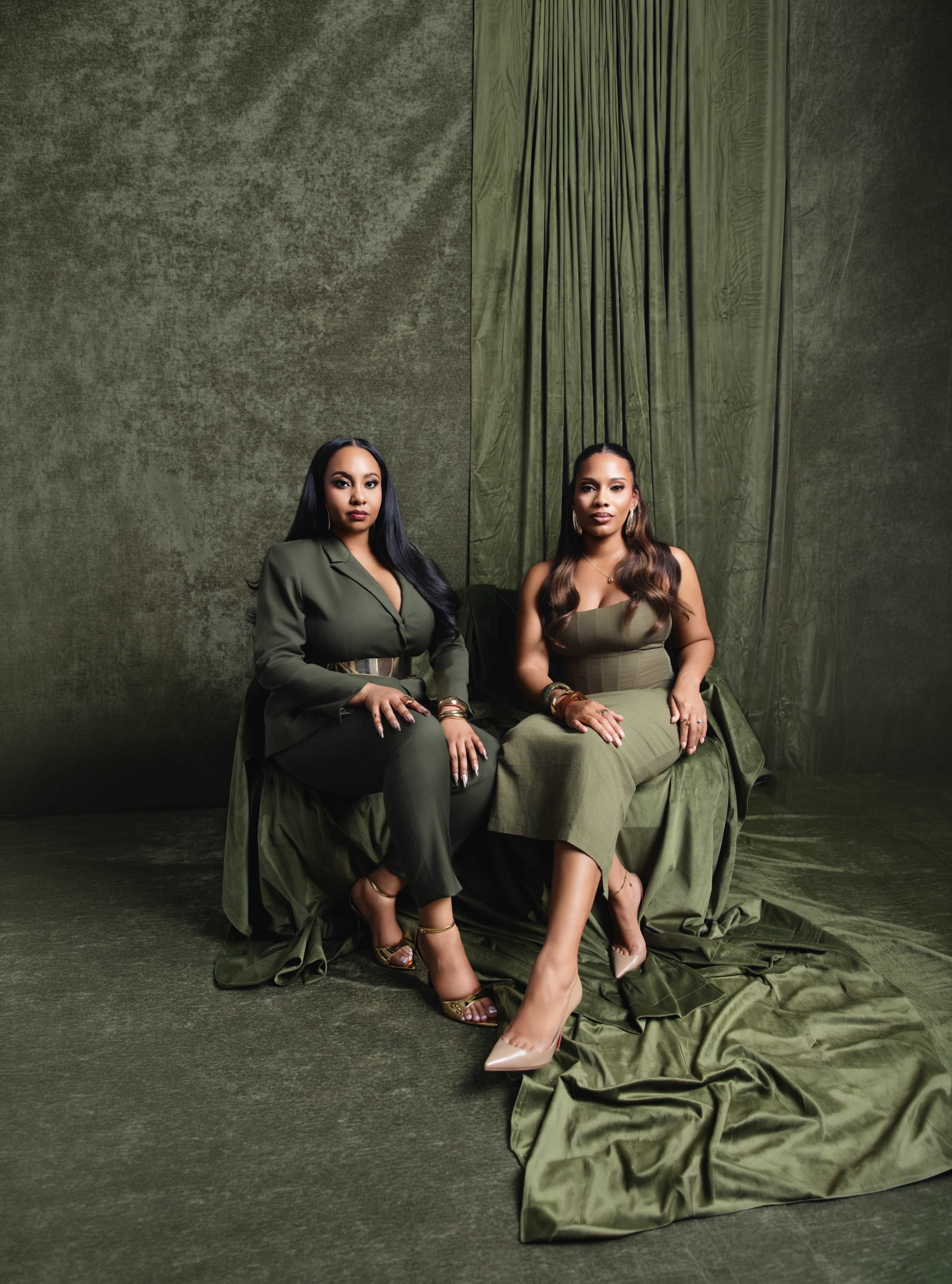 Two women are seated side by side on a green fabric backdrop, dressed in coordinated green outfits.