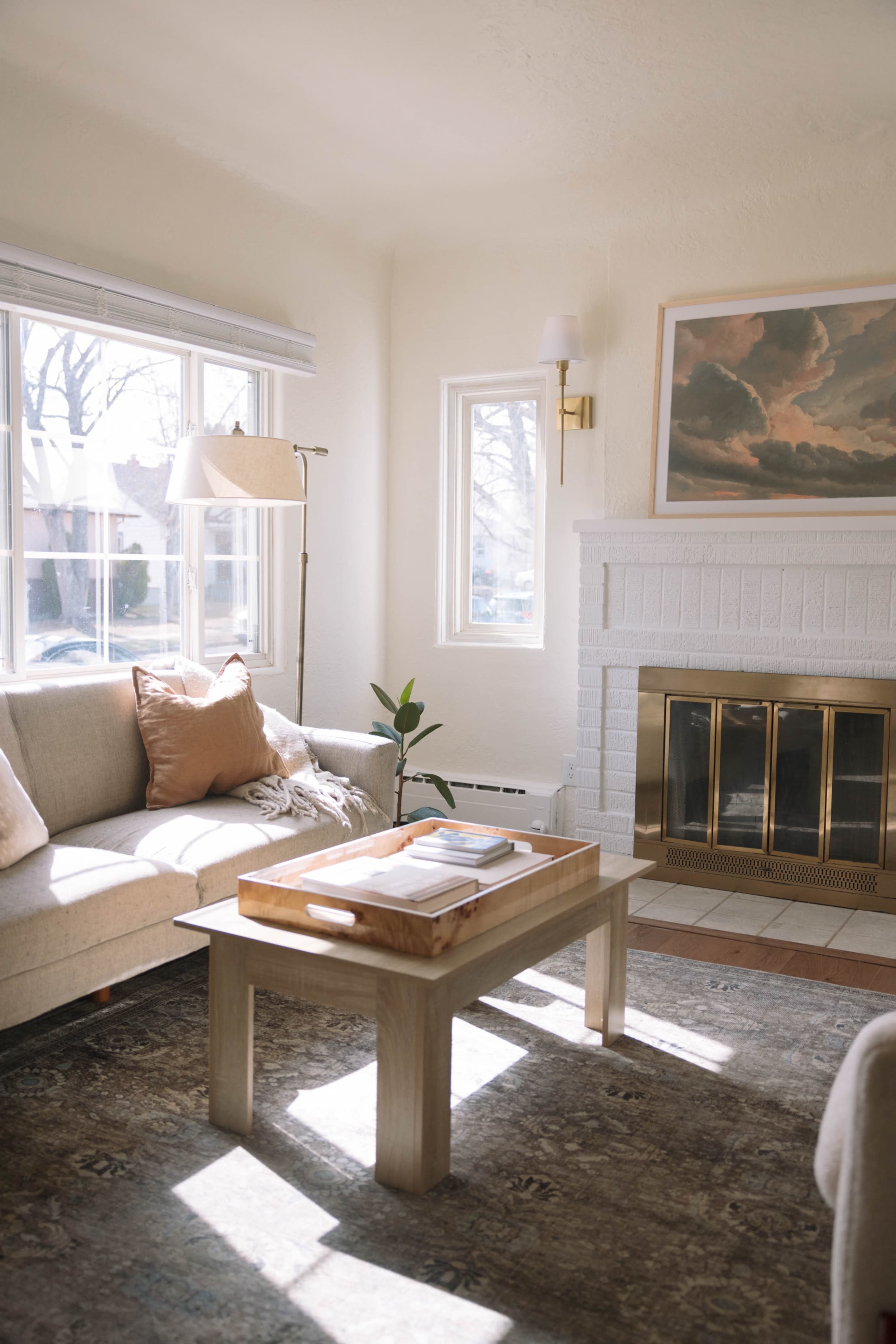 The image shows a bright living room with a beige sofa, a wooden coffee table, and a fireplace, illuminated by natural light from large windows.