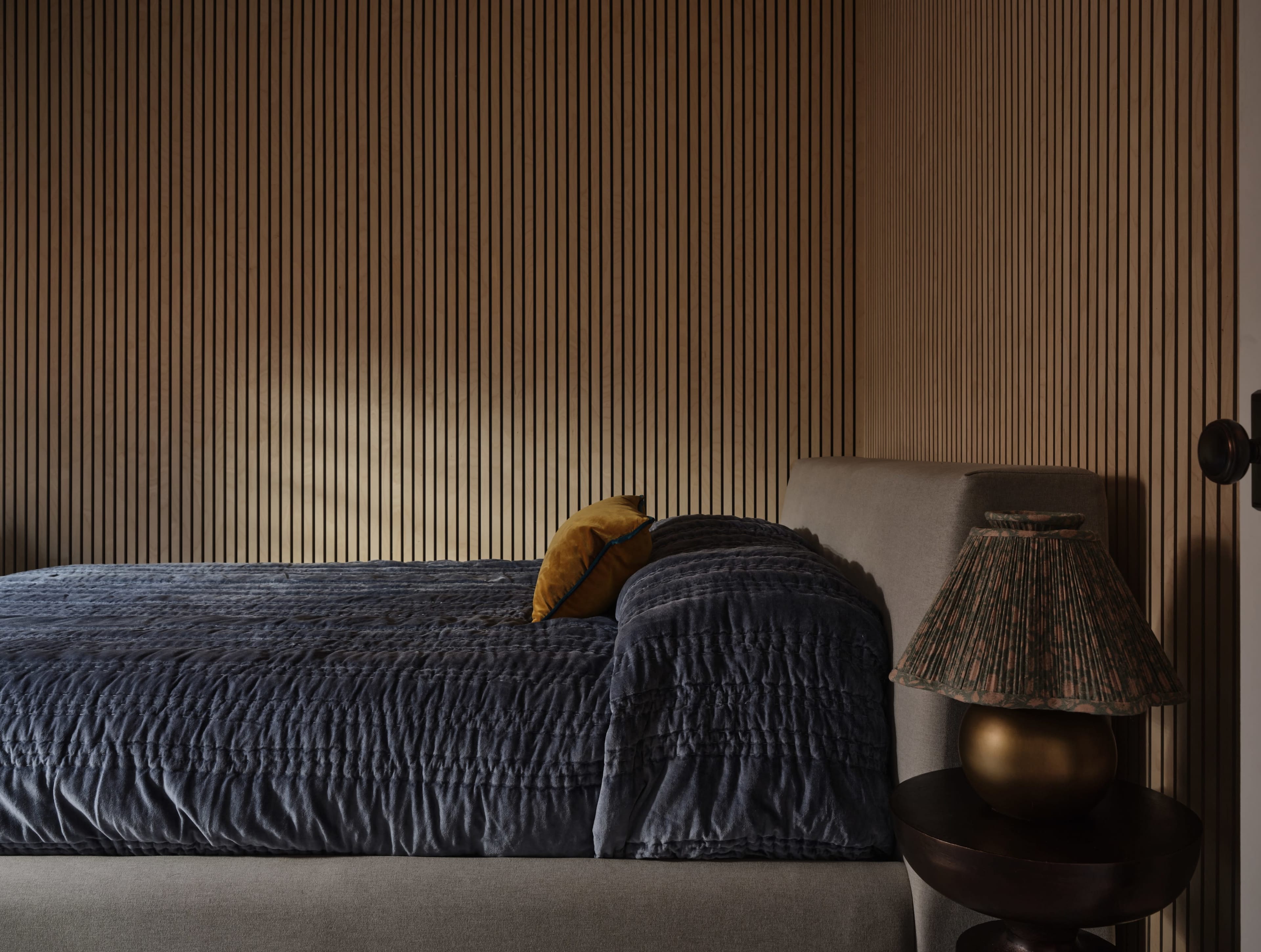 A neatly made bed with a textured dark quilt and a yellow cushion is positioned against a vertical-lined wooden wall, accompanied by a small table featuring a lamp with a patterned shade.