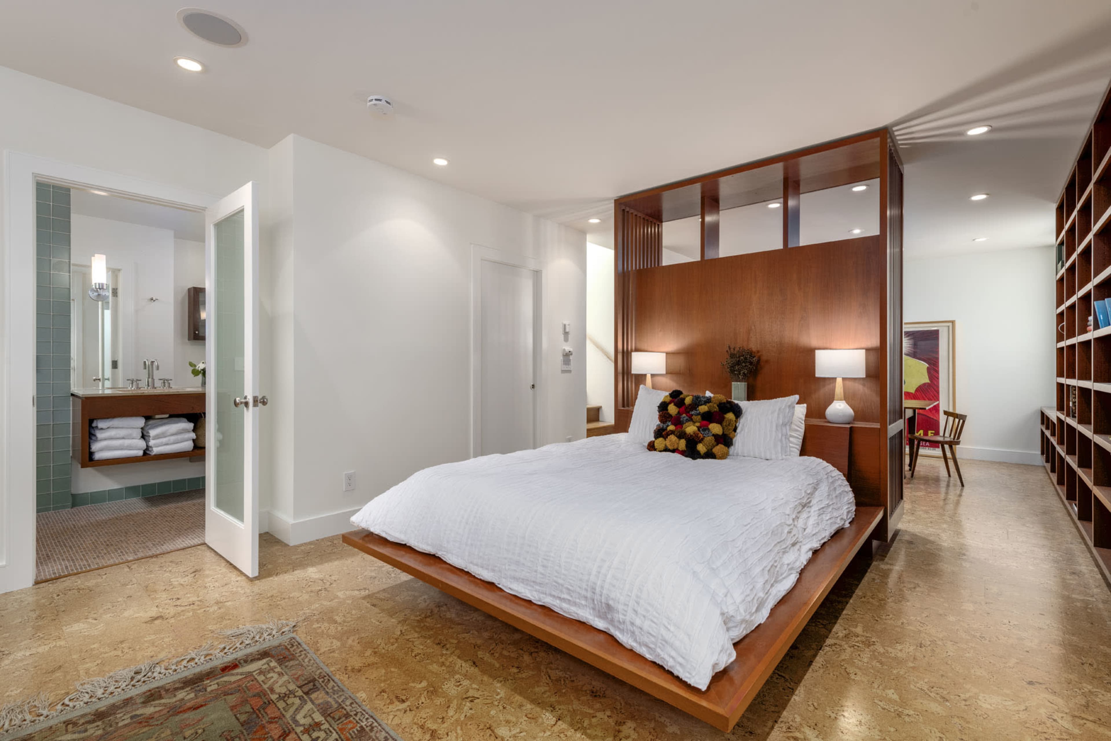 A modern bedroom features a wooden platform bed with a simple headboard, flanked by two lamps, and a view of a bathroom with green tiles and neatly folded towels.