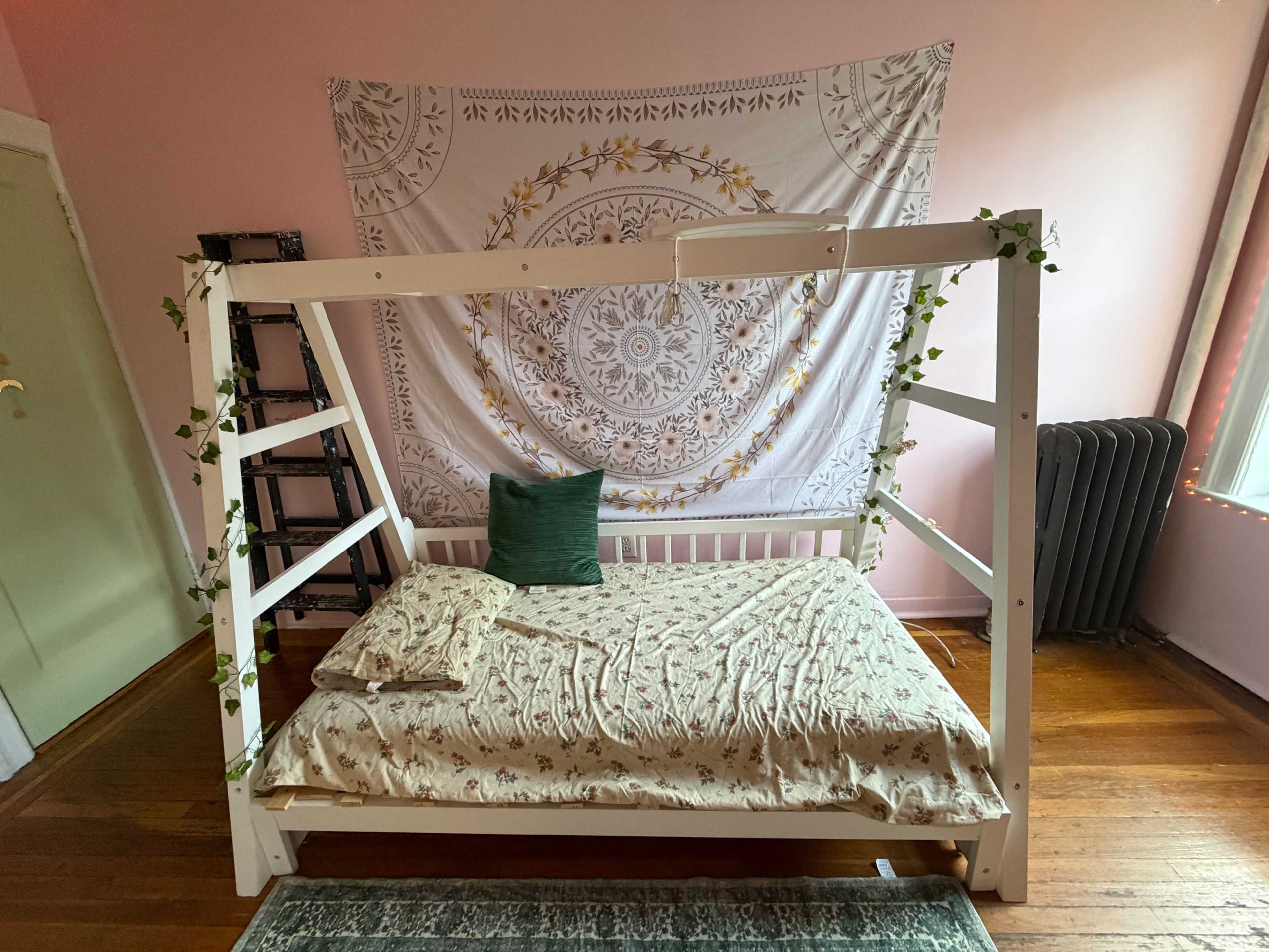 The image shows a minimalist bed frame with a patterned bedspread and a green pillow, set against a pink wall adorned with a decorative tapestry.