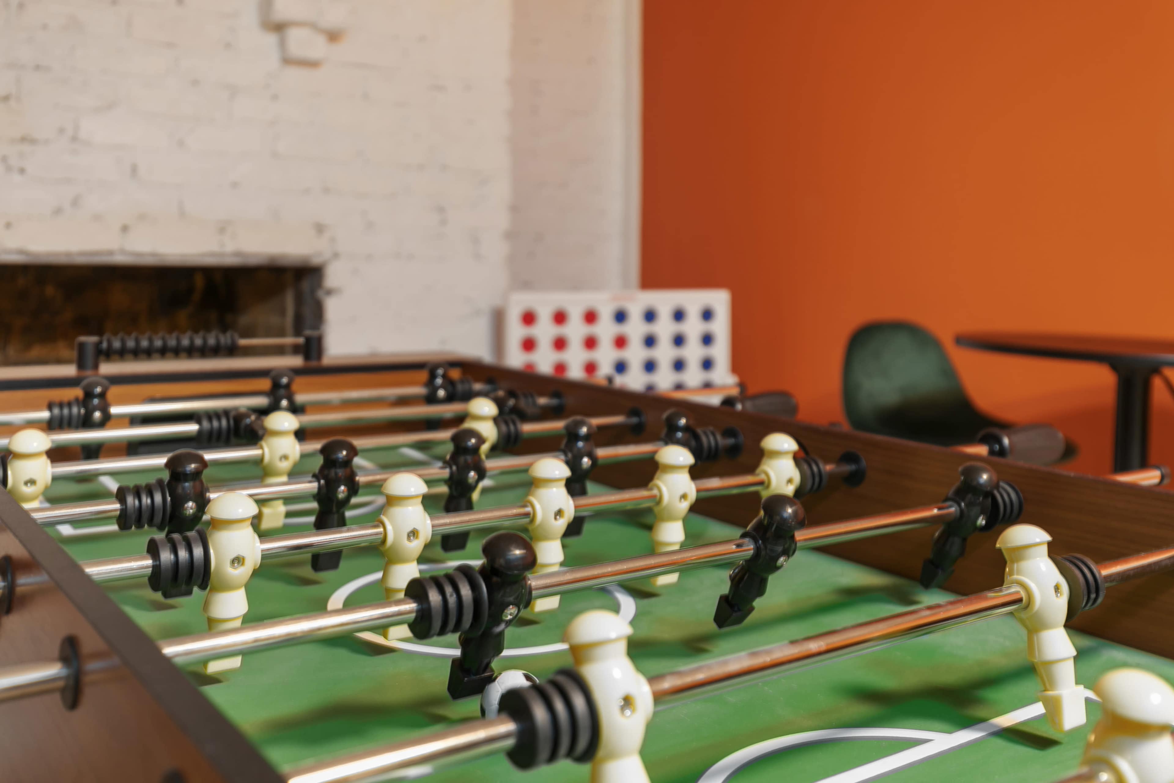 A foosball table with black and white figures is positioned in a room with an orange wall and a faint background of a Connect Four game.