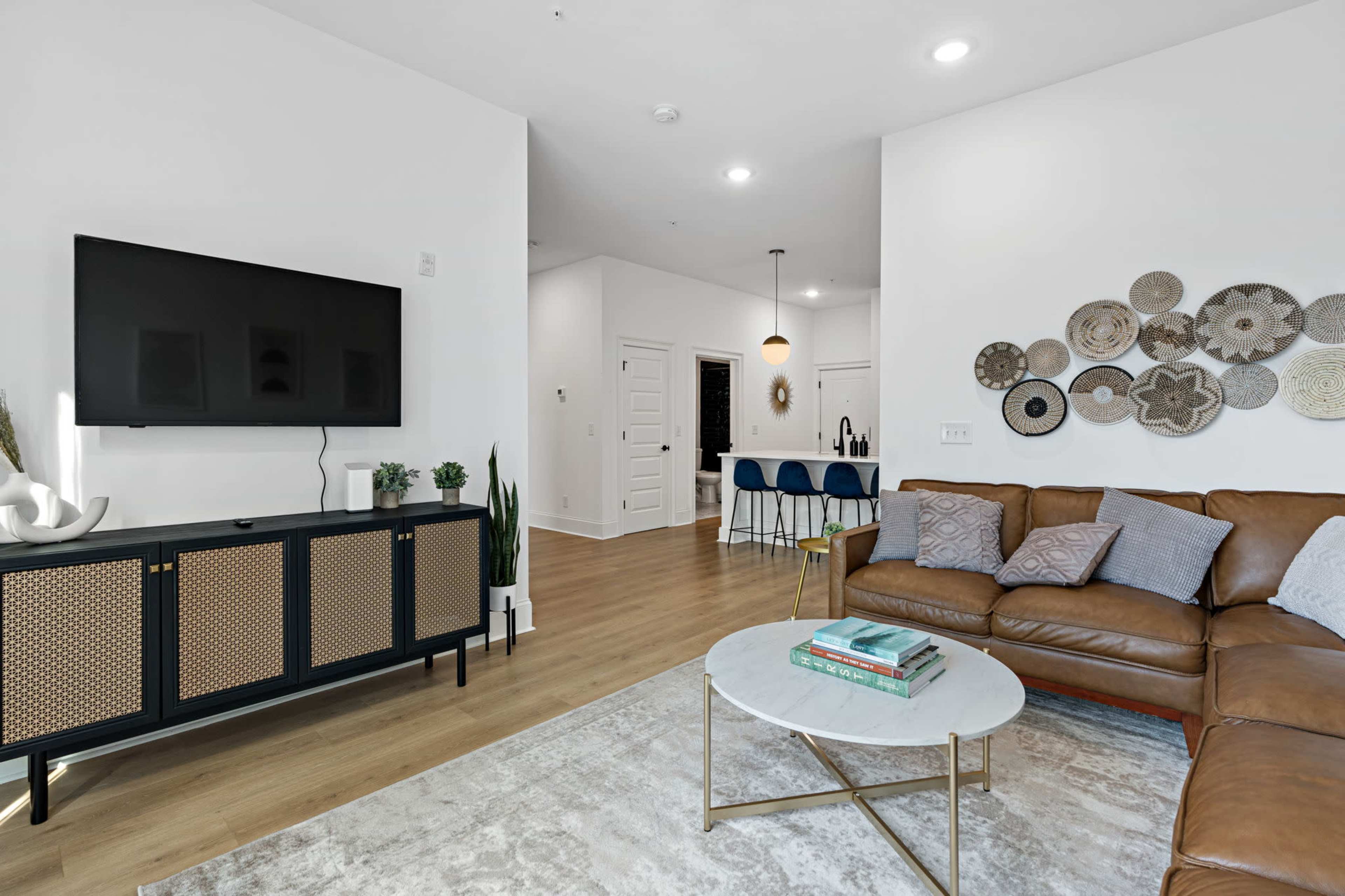 The image shows a modern living room with a brown sectional sofa, a circular coffee table, a wall-mounted TV, and a dining area in the background featuring bar stools and decorative plates on the wall.