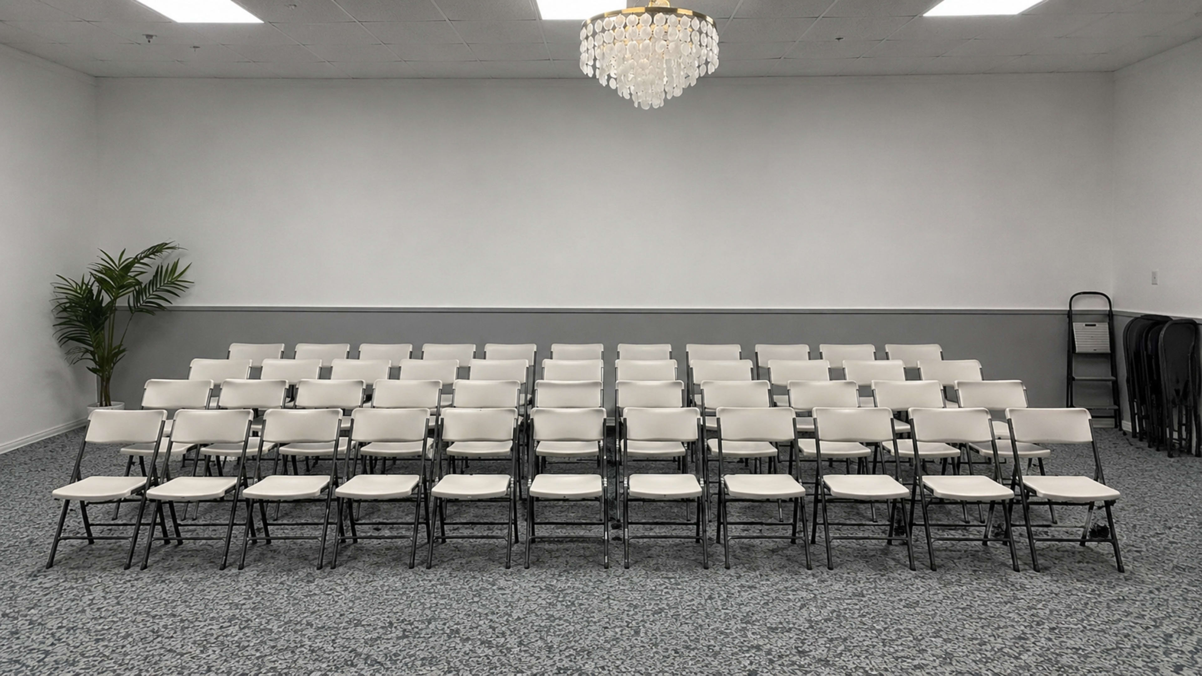 The image shows a neatly arranged set of black folding chairs positioned in rows in a spacious room with a chandelier and a decorative plant.