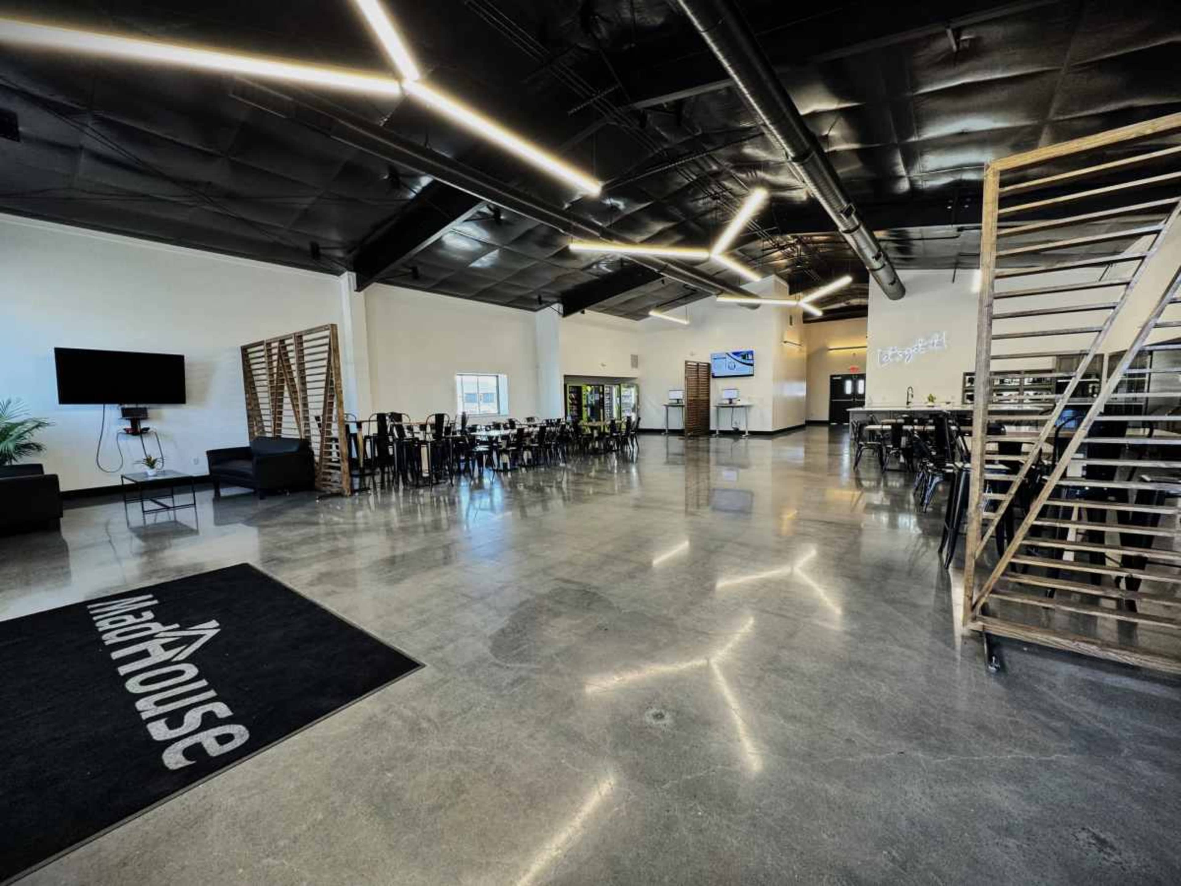 A spacious, modern interior of a building featuring polished concrete flooring, a large dining area with tables, a lounge area, and a staircase made of wood and metal.