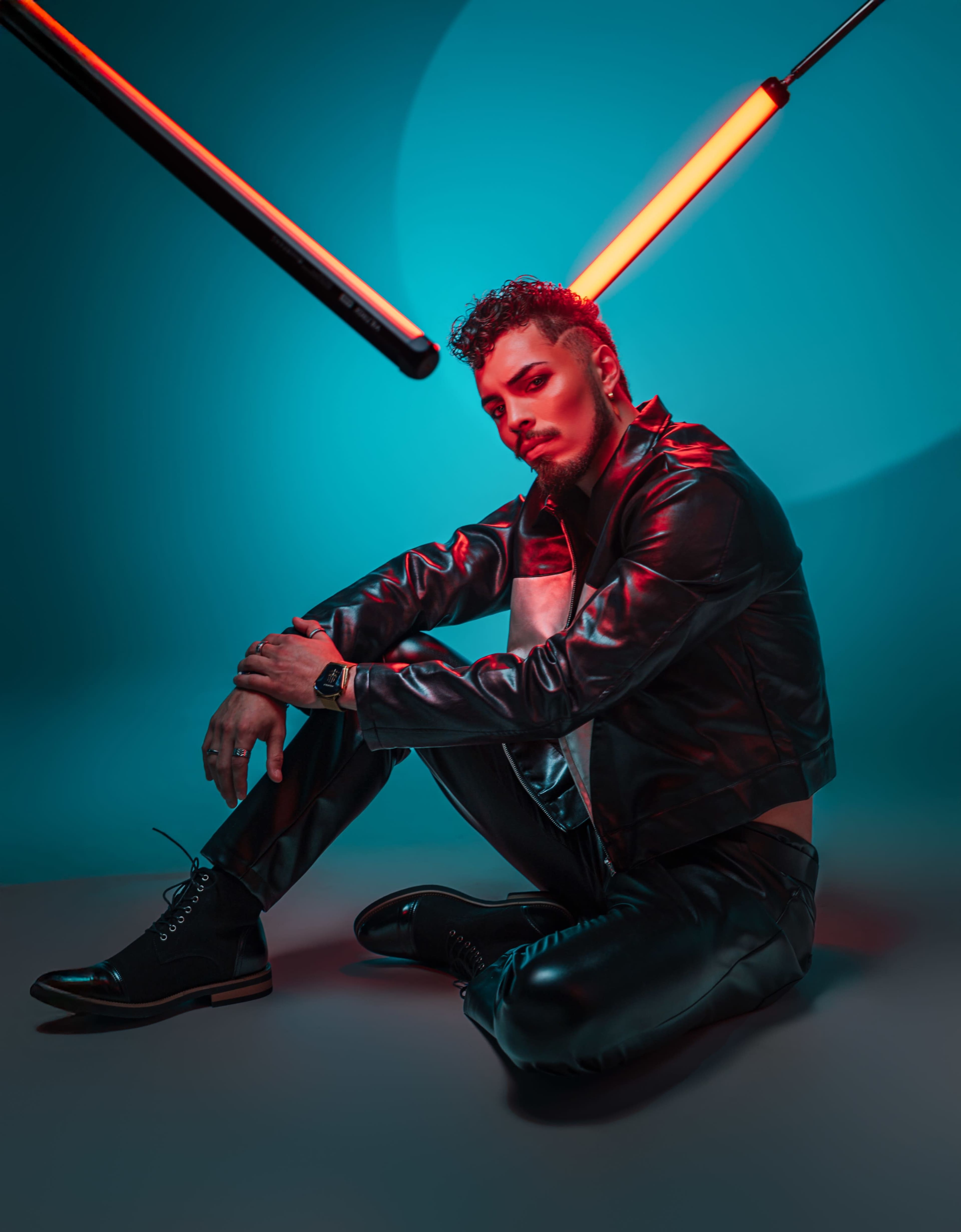 A person in a black leather jacket and pants sits on the ground under a glowing red light in a blue-toned environment.
