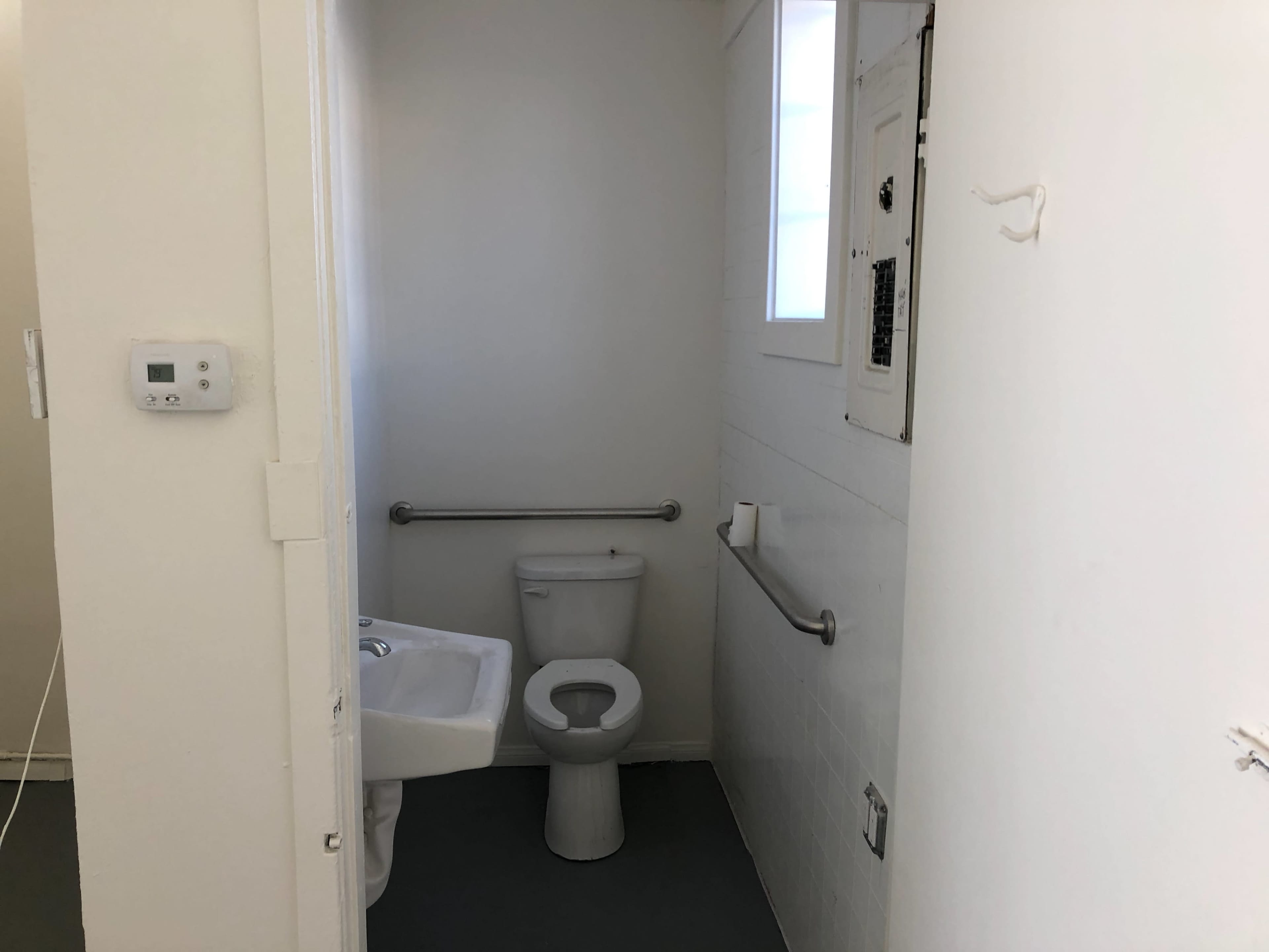 The image shows a small, clean restroom with a toilet, sink, and wall-mounted grab bars.