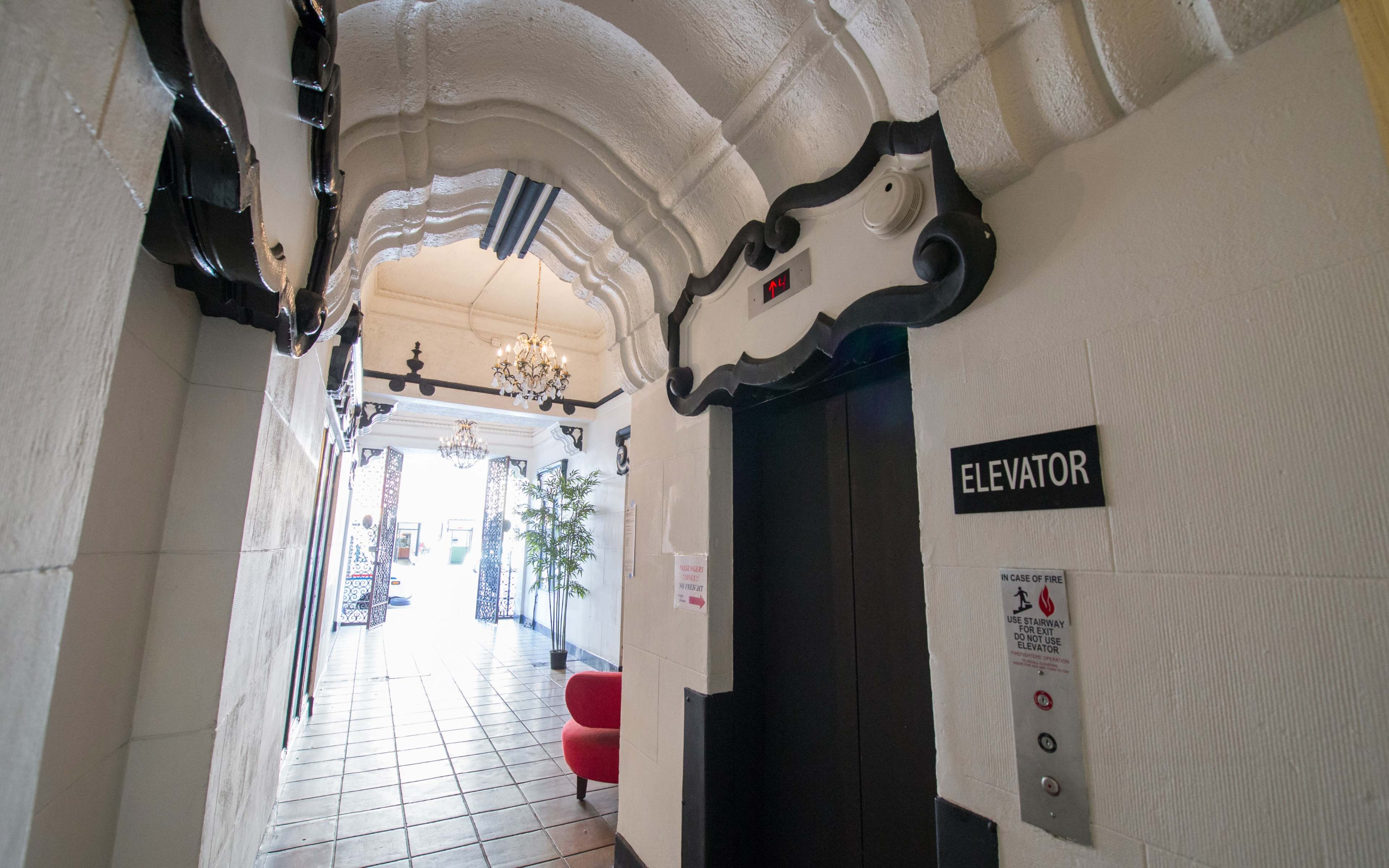 A corridor with an ornate ceiling, an elevator door, and an exit leading to a brightly lit area with a red chair and decorative elements.