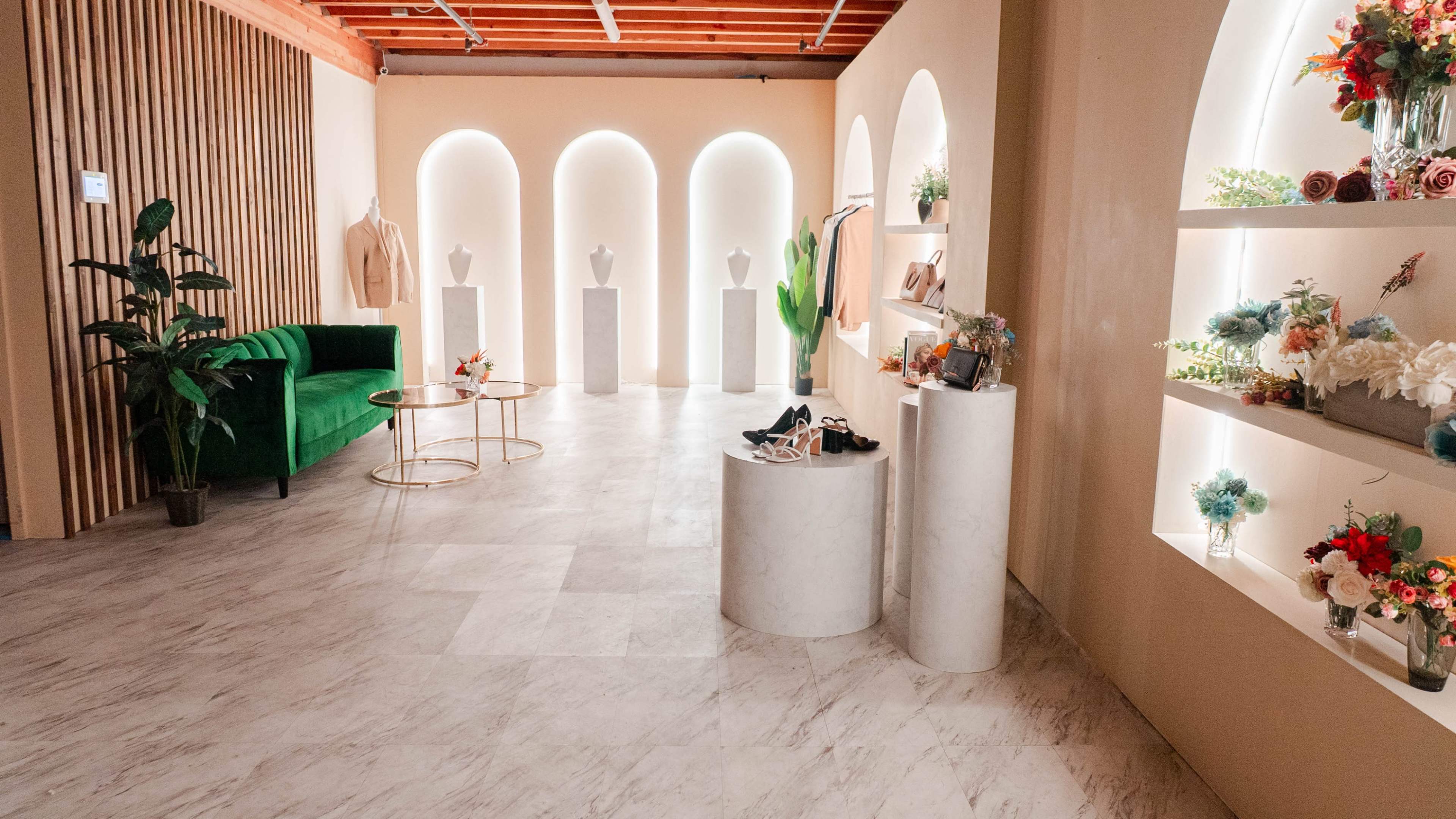 The image shows a stylish boutique interior featuring a green couch, marble display pedestals, and illuminated shelving showcasing various fashion items and floral arrangements.